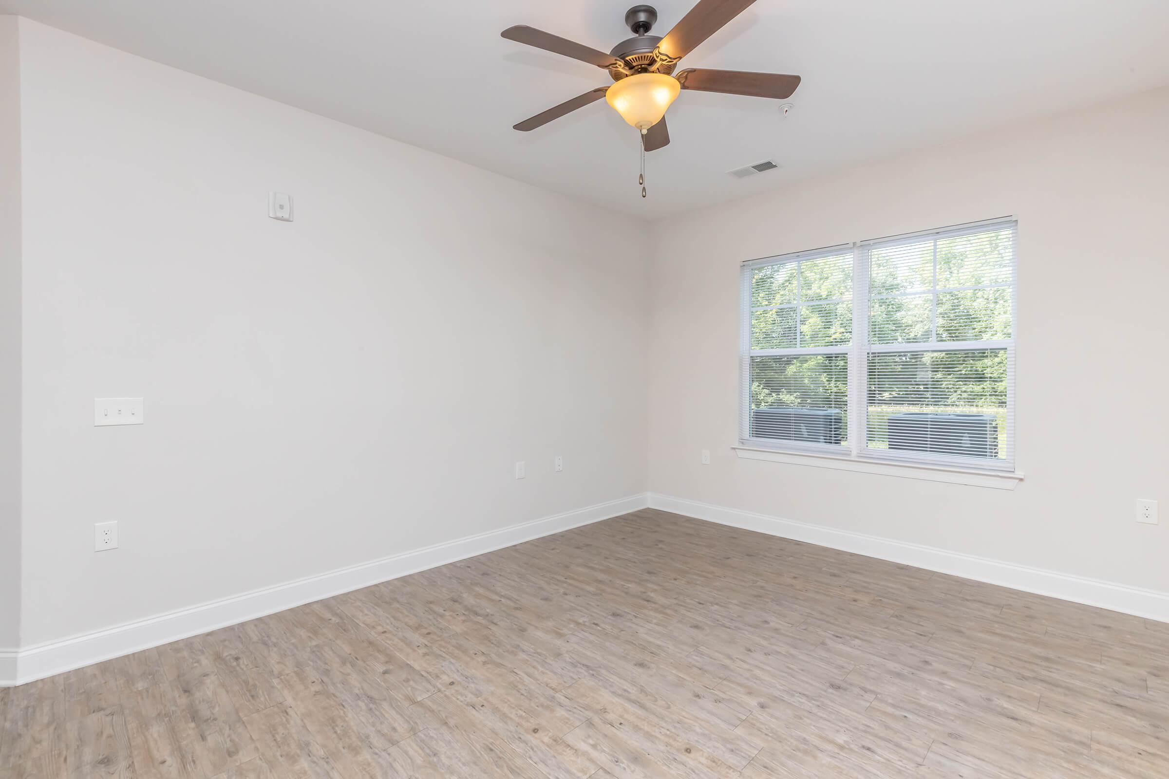 Spacious empty room with light-colored walls and a wooden floor. A ceiling fan with a light fixture hangs from the ceiling. There are two windows letting in natural light, framed by white trim, providing a view of greenery outside. The room is uncluttered, showcasing its potential for furniture arrangement.