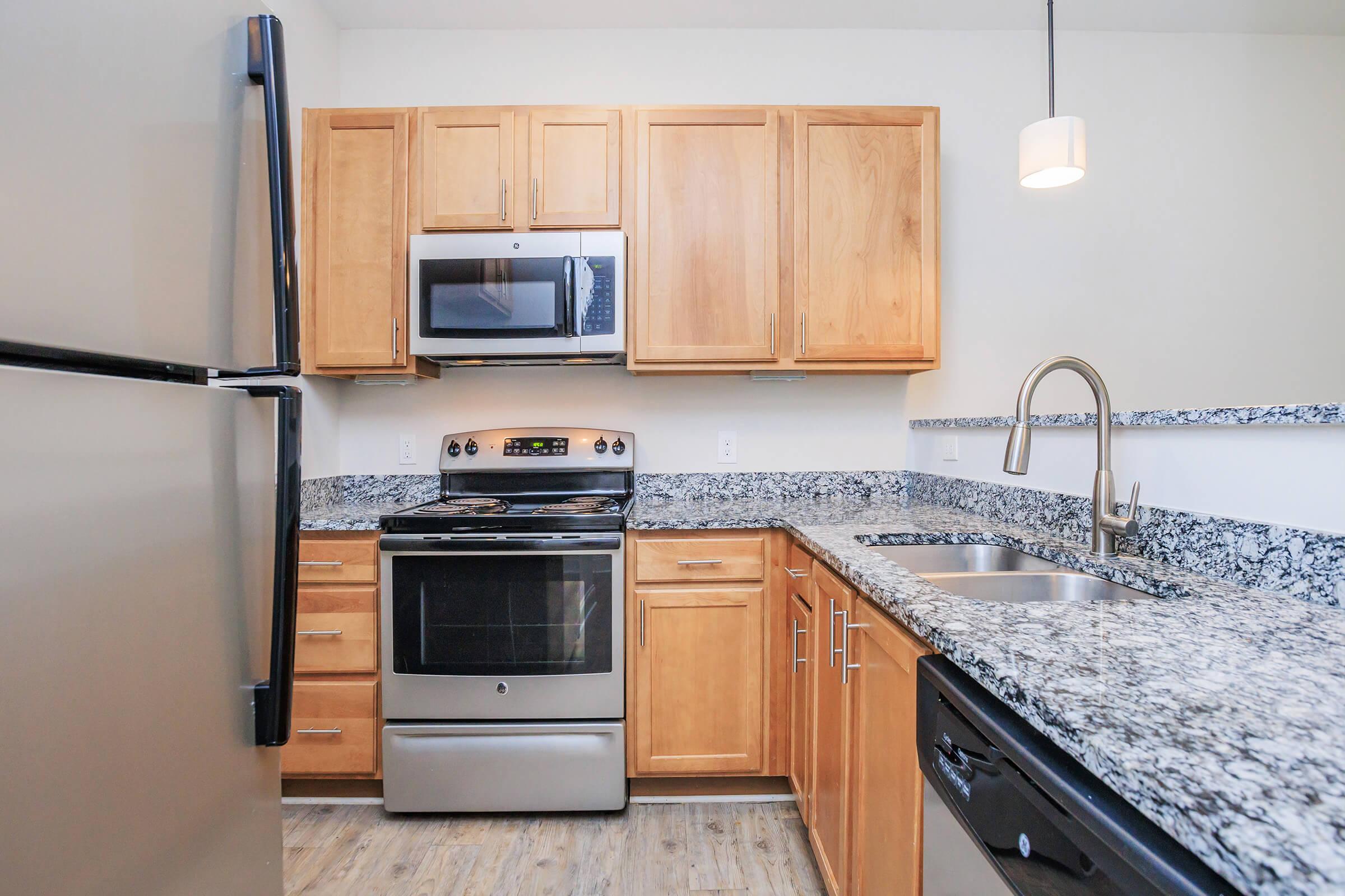 Modern kitchen featuring stainless steel appliances, including a stove and microwave, wooden cabinets, and granite countertops. There is a sink with a modern faucet, and an adjacent refrigerator. The design is clean and contemporary, with a light fixture above the countertop area.
