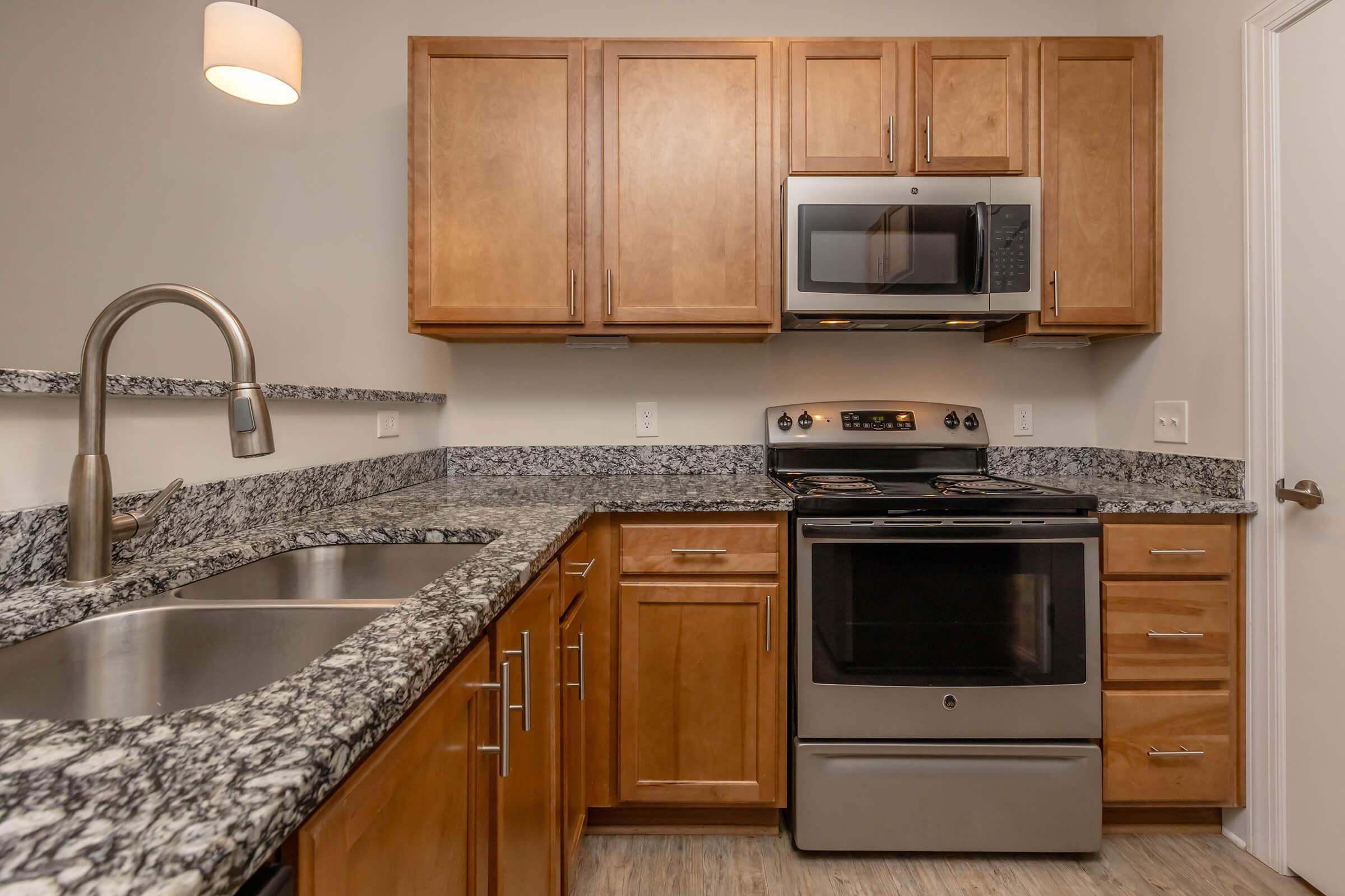 Modern kitchen featuring wooden cabinets, a granite countertop, stainless steel appliances including a microwave and oven, double sink, and a light fixture. The design is clean and functional, highlighting a contemporary style with warm tones.