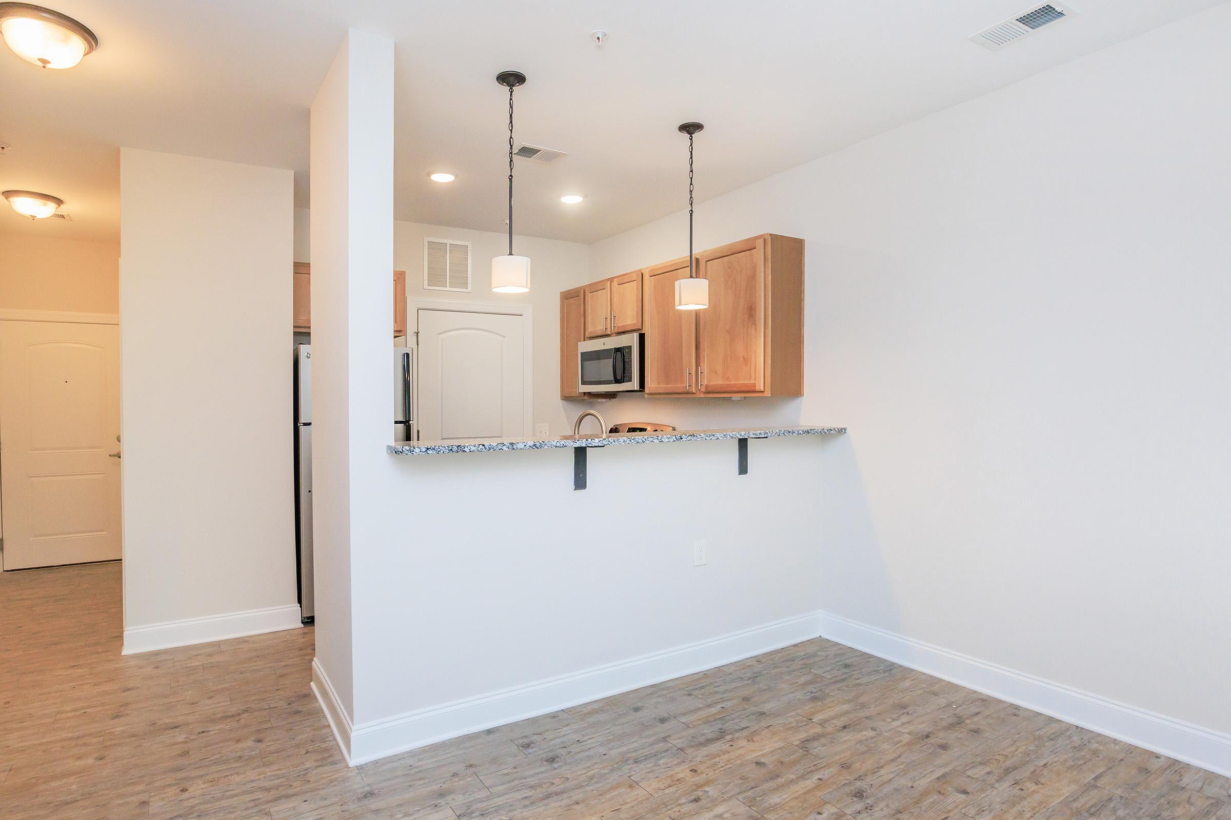 A modern kitchen and dining area featuring light wood cabinets, stainless steel appliances, and a breakfast bar with pendant lighting. The space has light-colored walls and wooden flooring, creating a warm and inviting atmosphere. A door leads to the entryway of the apartment.
