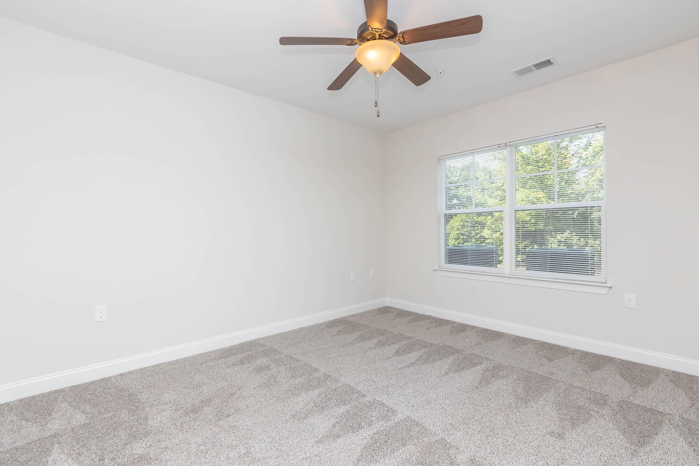 A spacious, empty room with light-colored walls and carpet. It features a ceiling fan with wooden blades and a window with blinds, allowing natural light to enter. The room is well-lit and ready for furnishing.