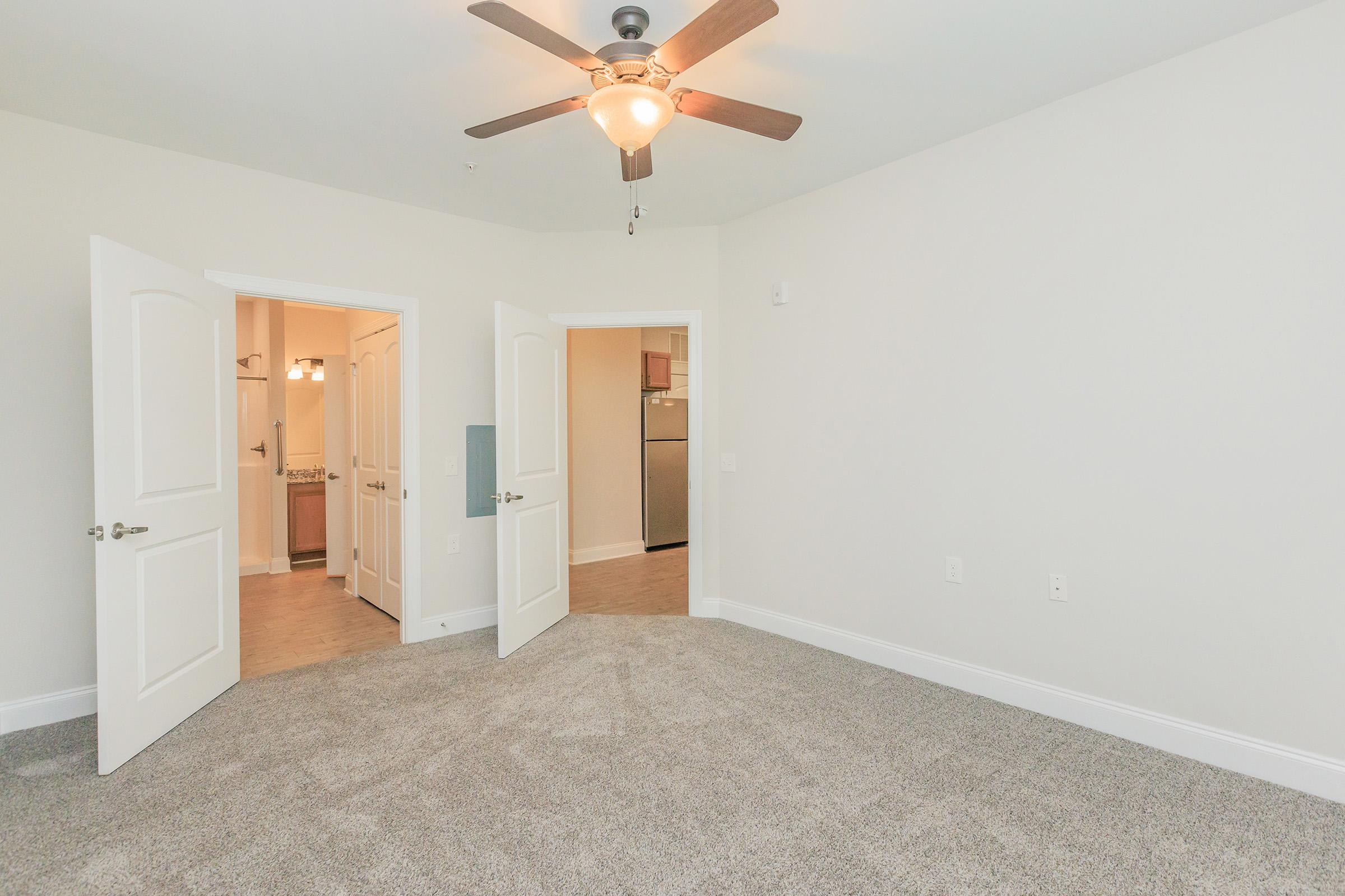 A spacious and empty room with light-colored walls and a ceiling fan. Double doors lead to another area, possibly a kitchen or bathroom. The floor is covered in plush carpet, and there is a warm, inviting atmosphere with ample natural light.