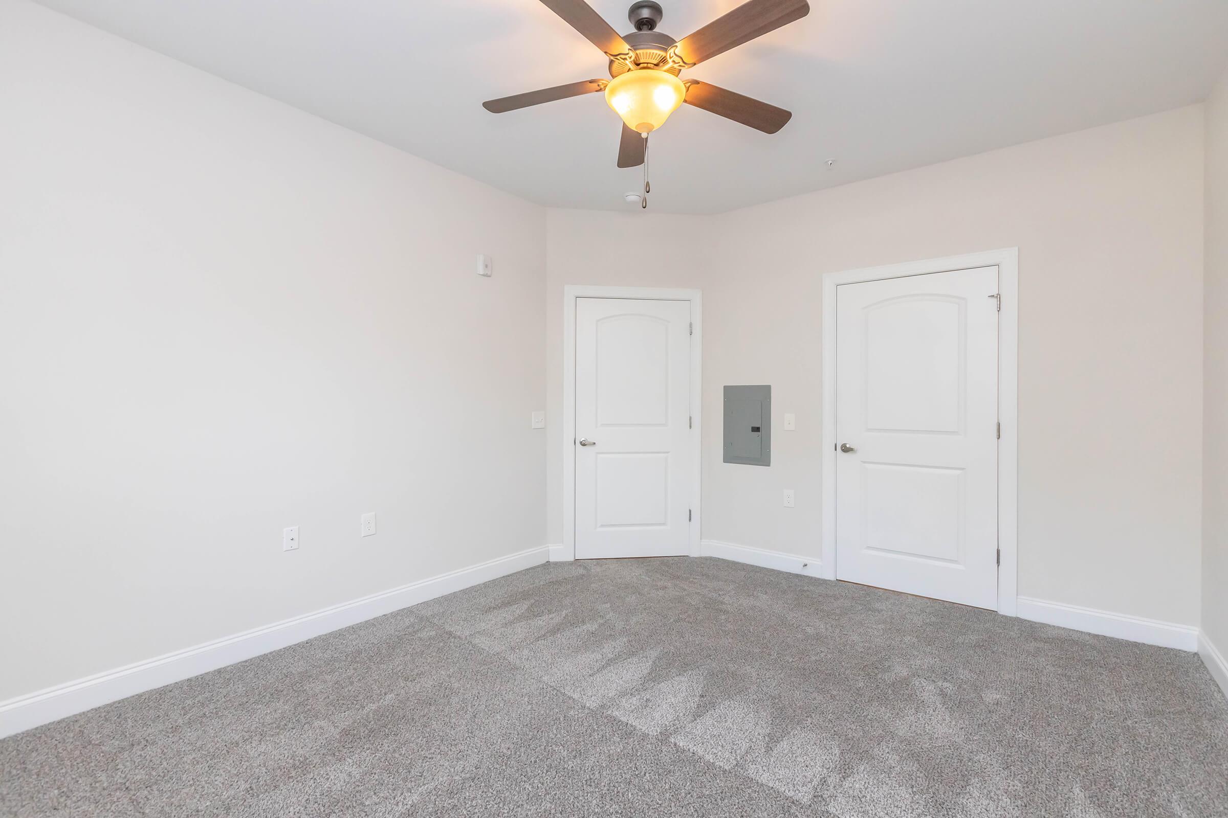 A spacious, empty bedroom featuring light gray carpet, a ceiling fan, and neutral-colored walls. Two closed doors are visible on the right, and a small wall-mounted unit is positioned near the door. Natural light brightens the room, emphasizing its airy and modern design.