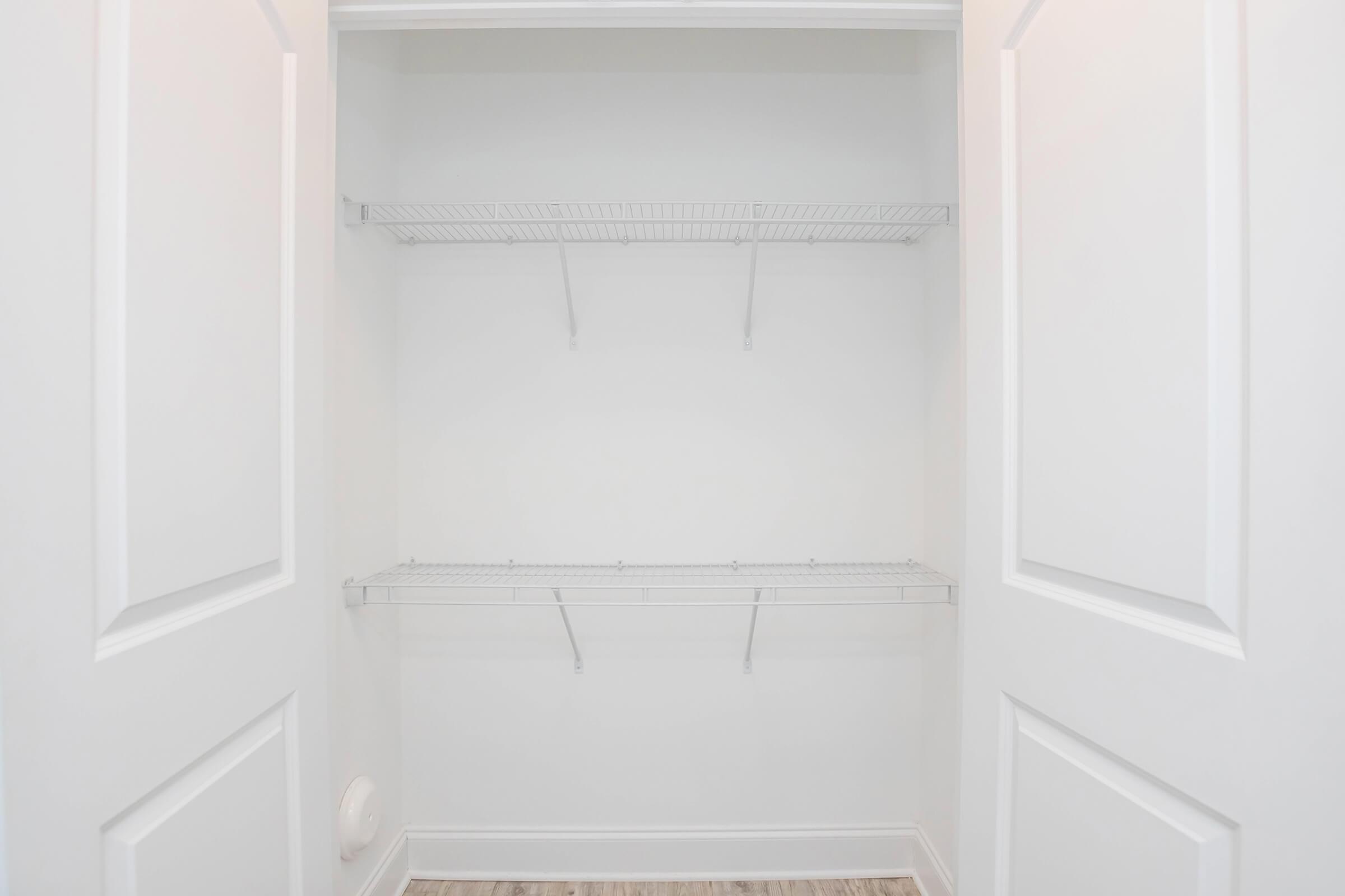 Empty closet with white walls and two wire shelving units. The closet doors are open, revealing a spacious interior with no items or decorations. The floor features light wooden planks, contributing to a clean and minimalistic appearance.