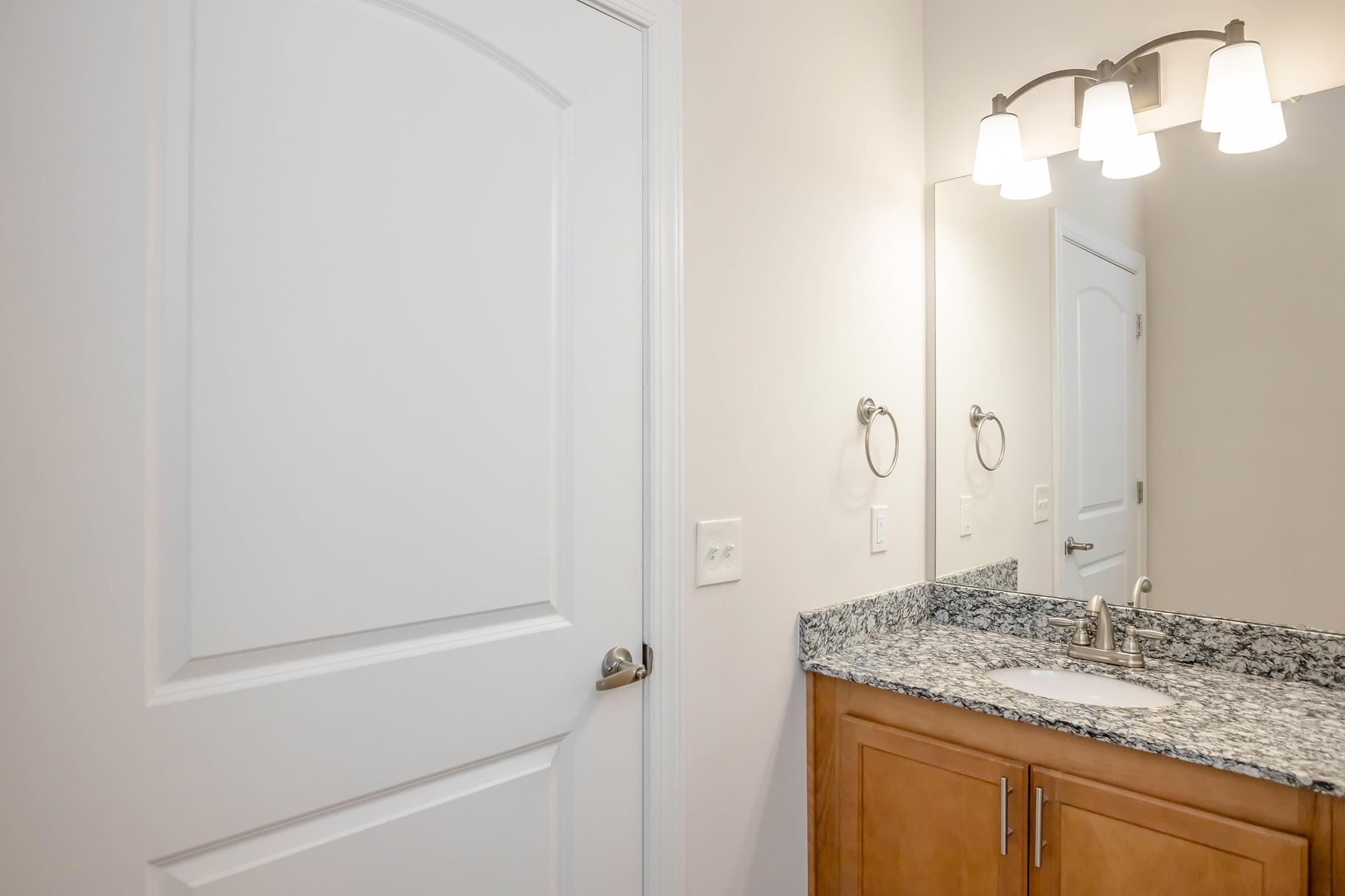 A modern bathroom with light-colored walls and a wooden vaniity. The countertop features grey and white granite, and there's a round sink. Above the sink, a wall-mounted mirror reflects a light fixture with three bulbs. A white door is visible on the left side of the image.