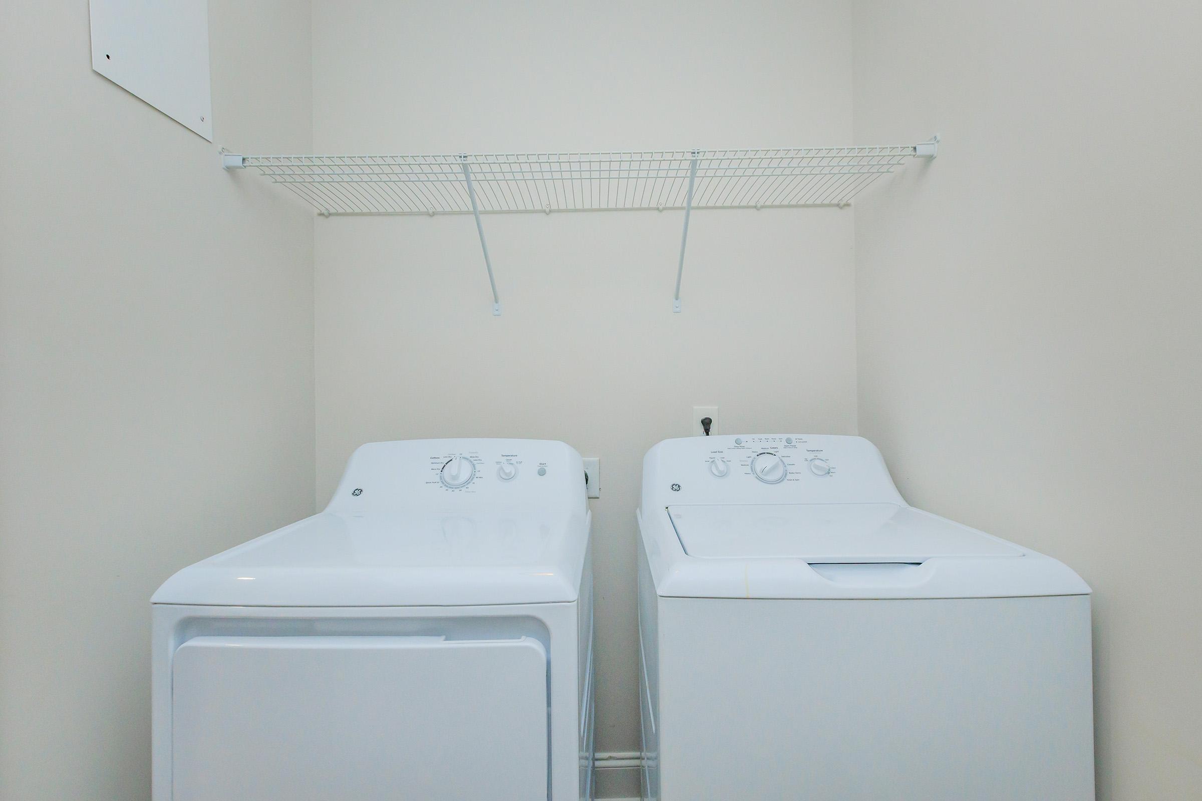Two white laundry appliances are situated side by side in a small, neutral-colored laundry room. A washing machine is on the left and a dryer is on the right. Above them, a wire shelf is mounted to the wall, providing space for storage. The walls are a light beige, creating a clean and simple atmosphere.