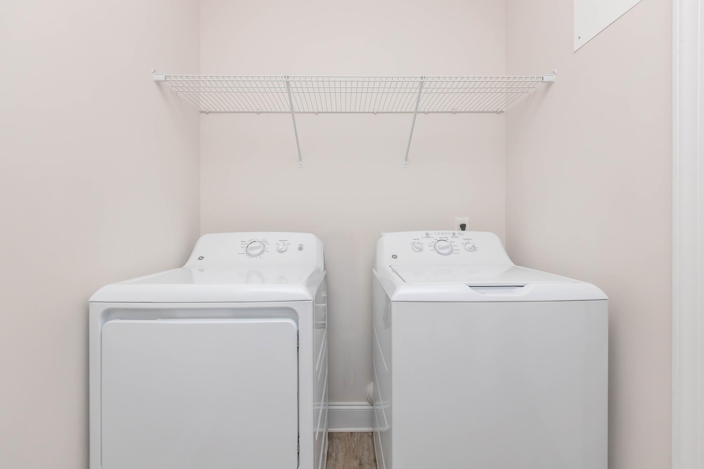 A clean laundry room featuring a white washing machine and a matching dryer. There is a wire shelf above the appliances for storage, and the walls are painted in a soft pink color, providing a bright and inviting atmosphere. The flooring is light-colored wood, enhancing the overall neat appearance.