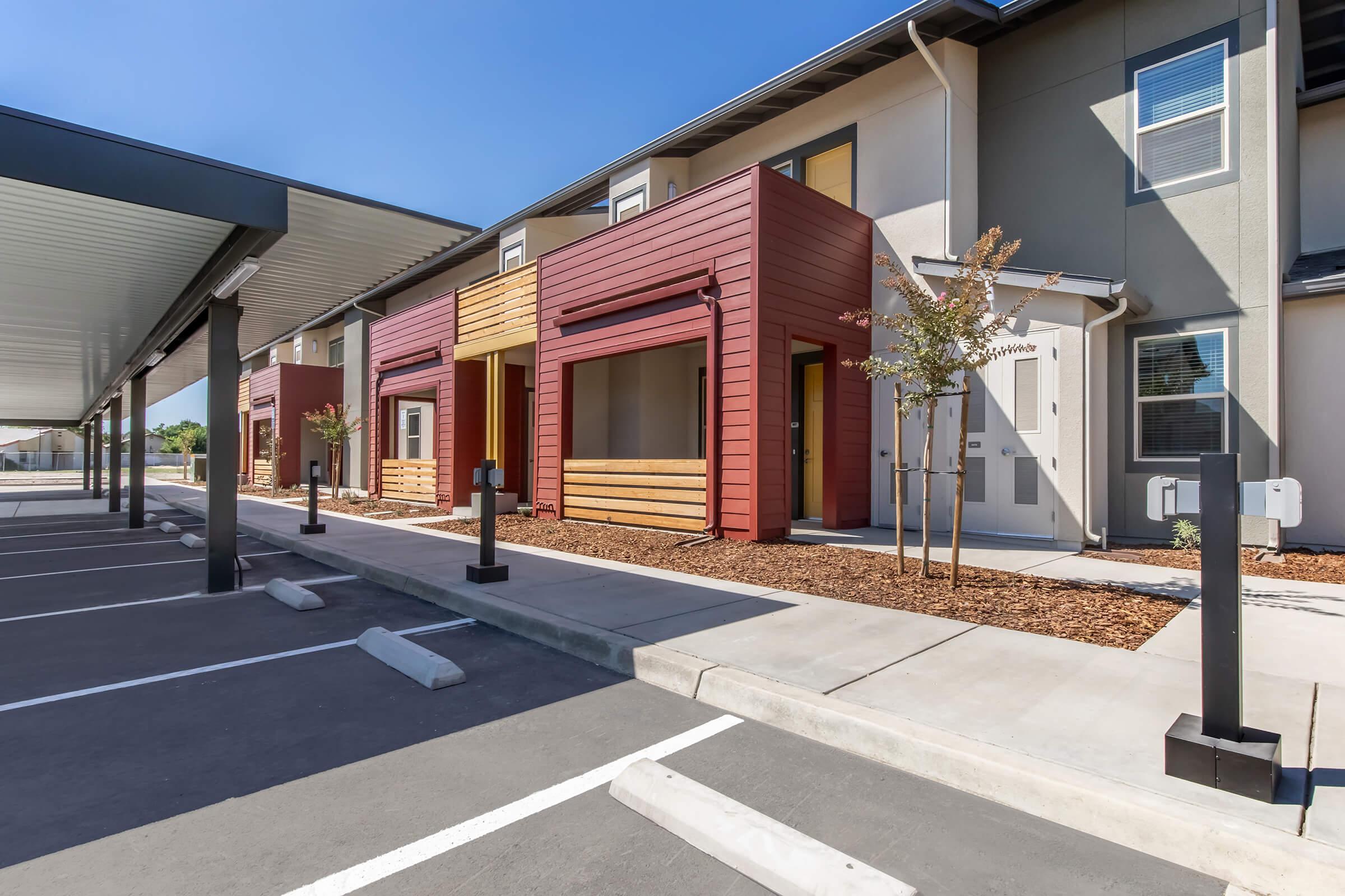 Modern multi-unit residential building with colorful exterior, featuring red and tan facades. A parking area in front with several empty spaces and lampposts, surrounded by clean landscaping and young trees. Clear blue sky enhances the bright, welcoming atmosphere of the property.
