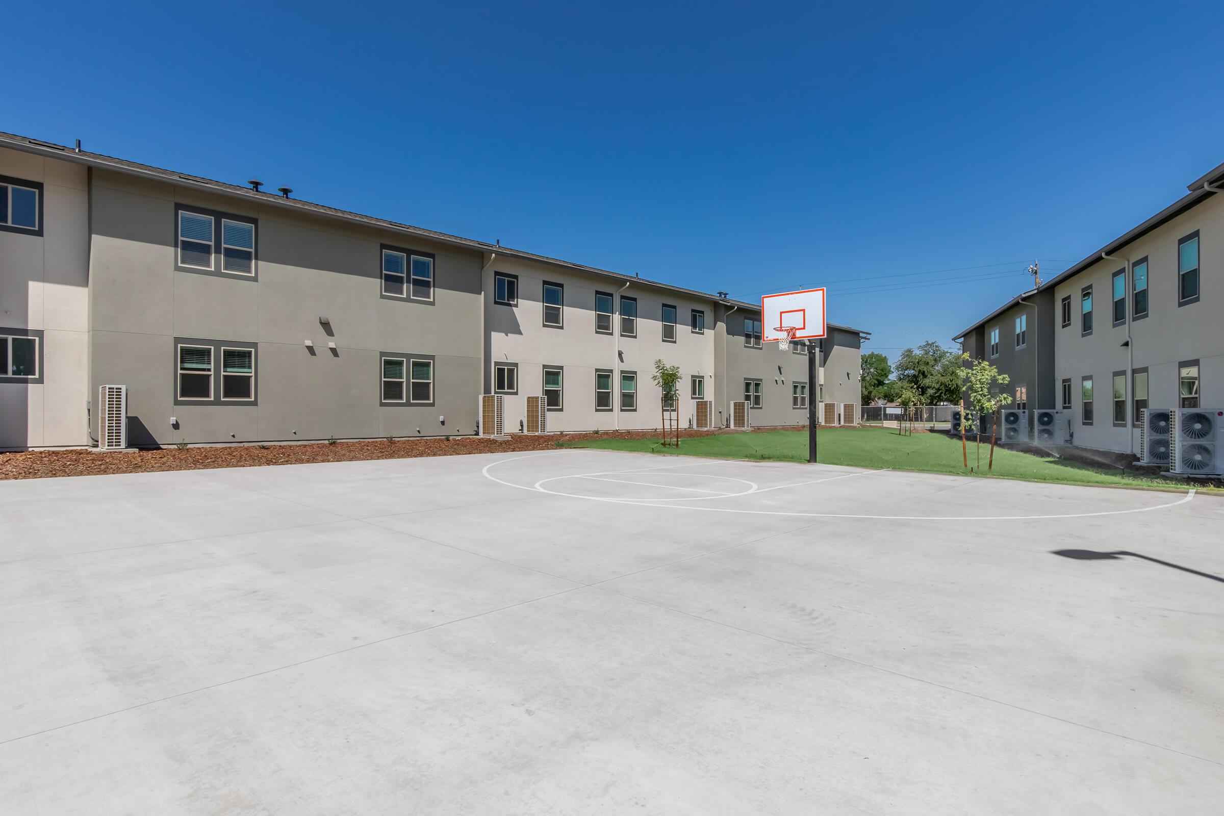 An outdoor basketball court featuring a hoop, surrounded by modern multi-story buildings. The court is situated in a sunny area with clear blue skies, and there are small trees and air conditioning units visible along the perimeter. The ground is made of concrete.