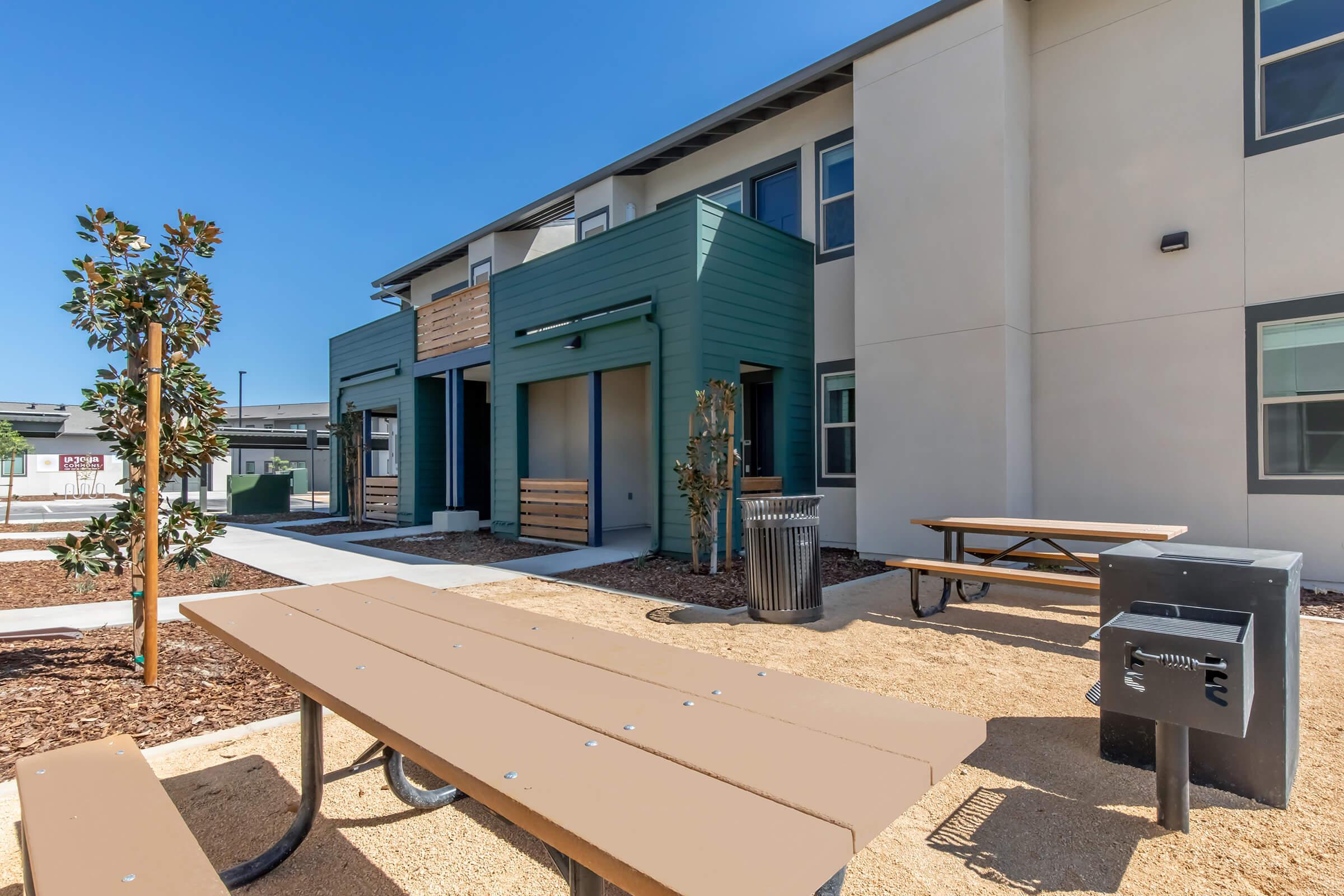 View of a modern commercial building featuring a green and beige exterior. The image includes a well-maintained outdoor area with picnic tables, a barbecue grill, and landscaping with small trees. There are clear skies above and a pathway leading to the entrances of the building.