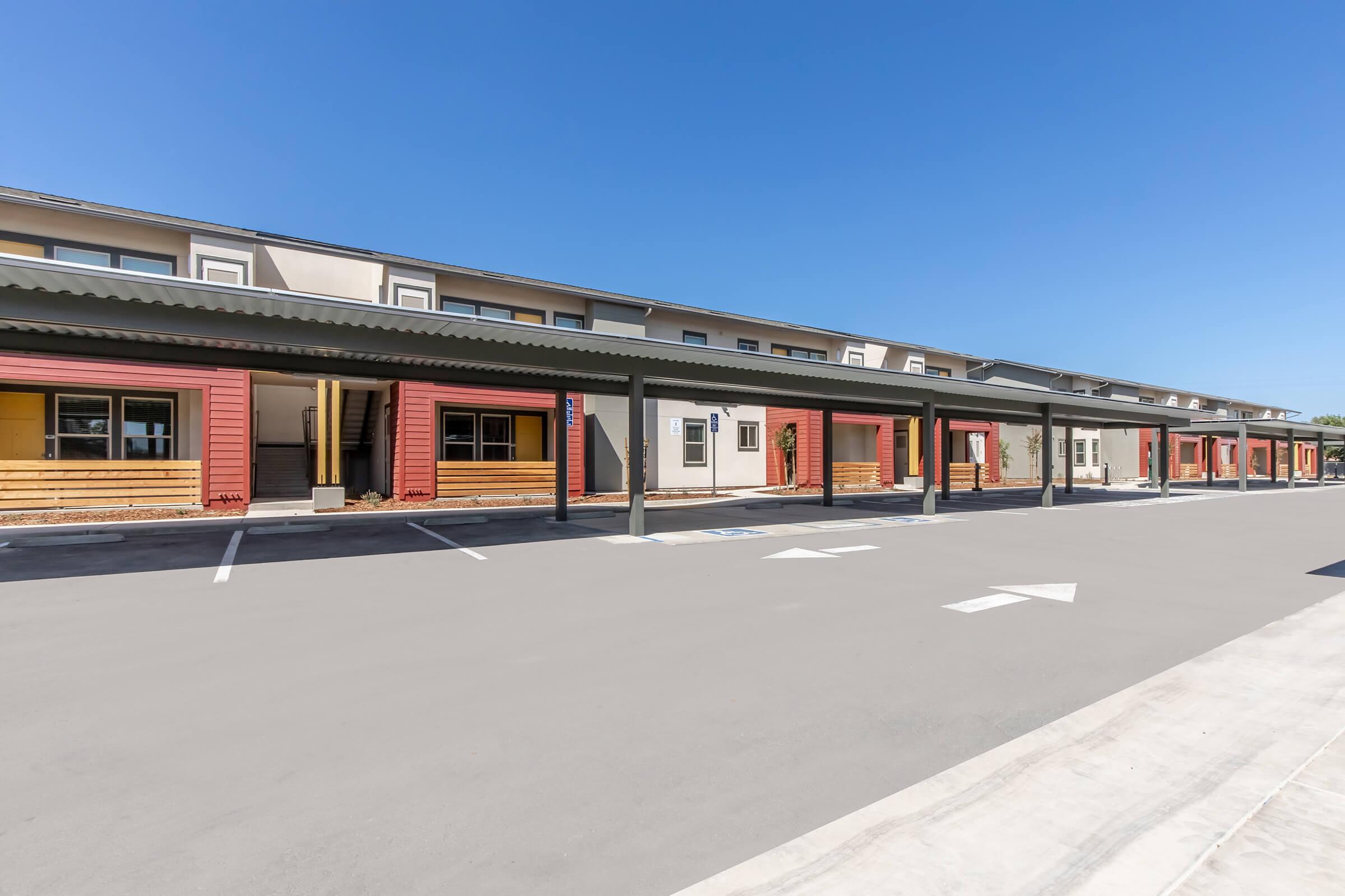 A modern, single-story building with a row of covered parking spaces in front. The building features a mix of vibrant stucco colors, including pink and beige, under a clear blue sky. Sidewalks lead to entrances, creating a welcoming atmosphere in a residential or commercial area.
