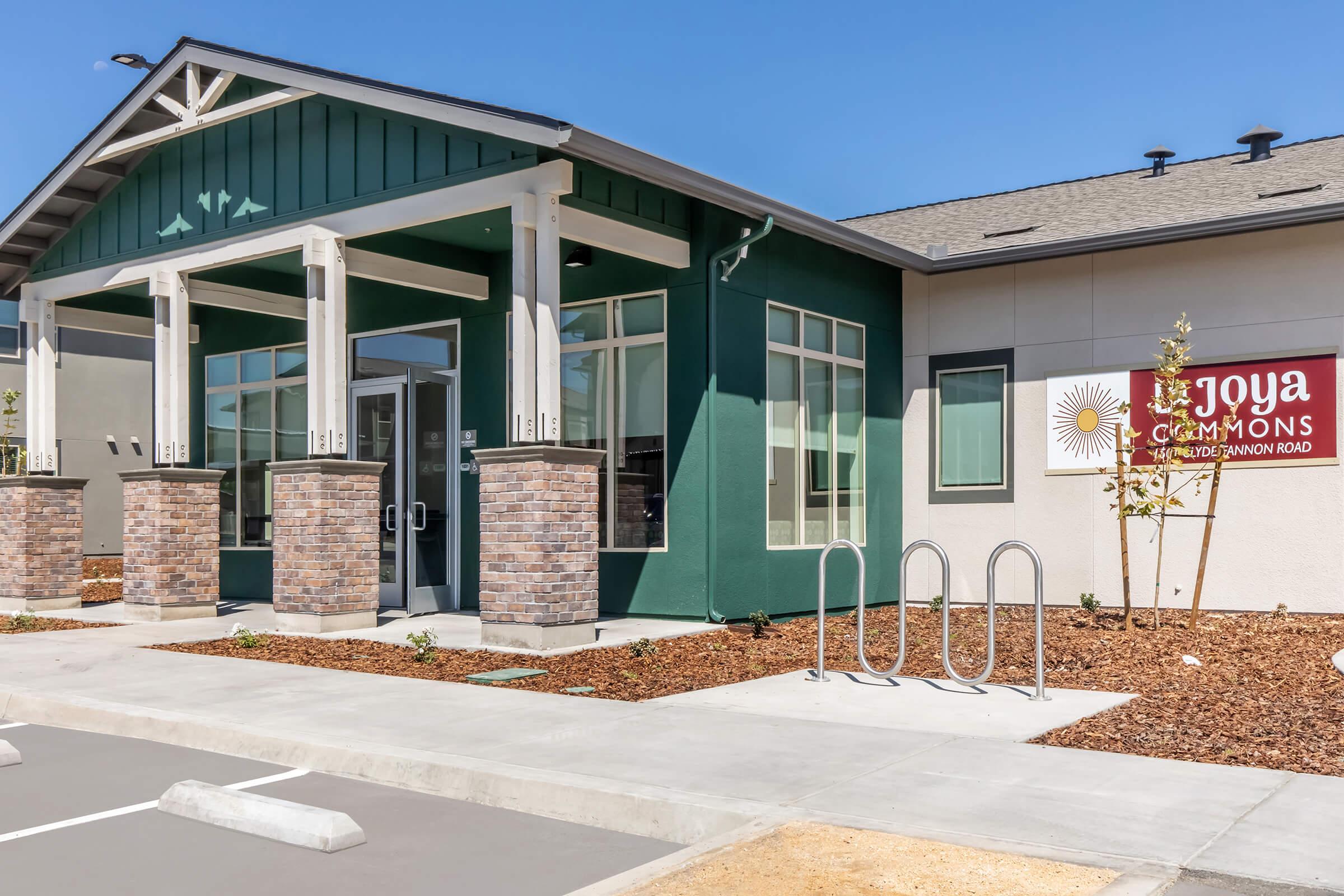 Exterior view of Joya Commons, a modern building featuring green and light-colored walls, decorative stone columns, and large windows. There is a sign displaying the name "Joya Commons" and a bike rack in front. The surrounding landscape is landscaped with gravel and small plants.