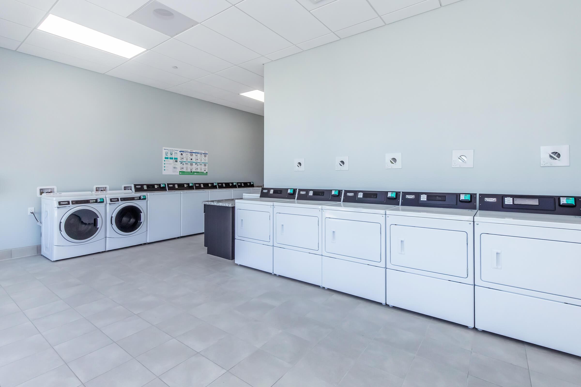 Bright and modern laundry room featuring several commercial washing machines and dryers along one wall. The machines are arranged neatly with a countertop and a wall-mounted information board in the background. The spacious room has gray tiled floors and bright overhead lighting.