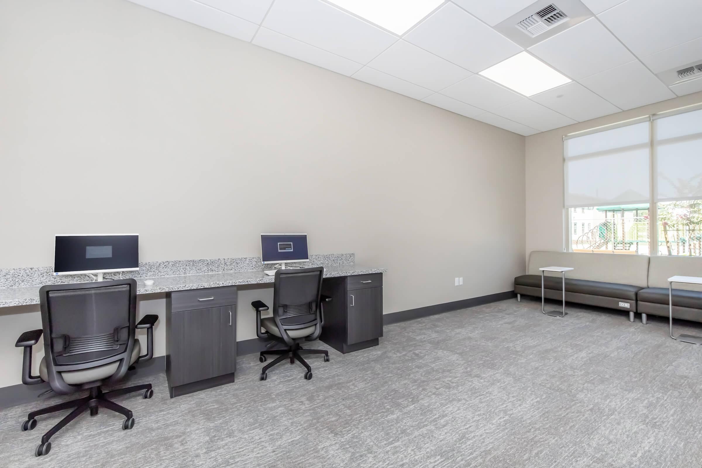 A modern, minimalistic office space featuring two workstations with desktop computers on gray counters. There are ergonomic black chairs at each workstation. The room has light gray walls, a carpeted floor, and large windows with blinds. A sleek gray couch is positioned against the wall, providing a comfortable seating area.