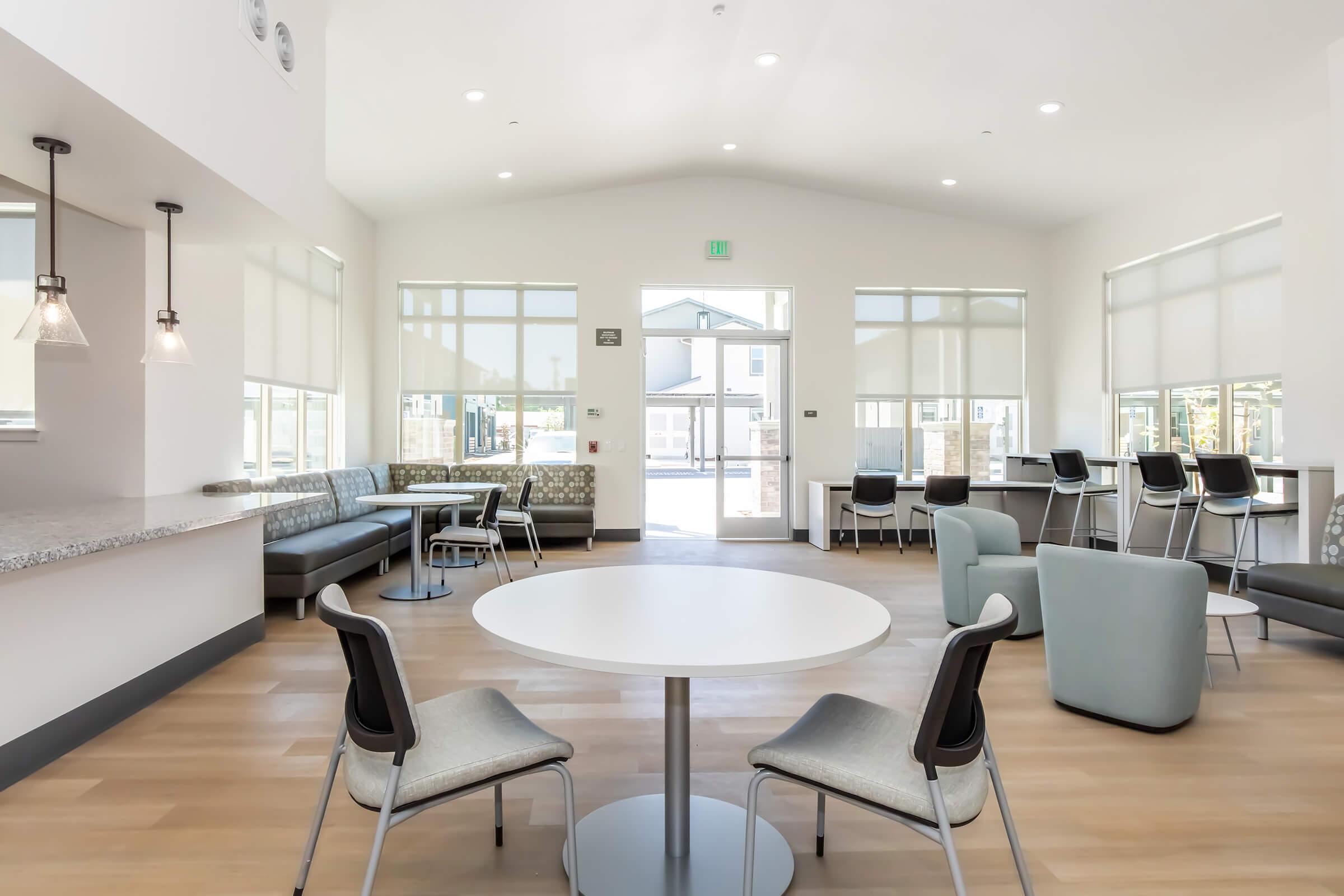 A modern, bright lounge area featuring a round table with chairs, a gray sectional couch, and high-top seating near the windows. Large windows allow natural light in, and the space has a clean, contemporary design with wooden flooring and minimal decor.