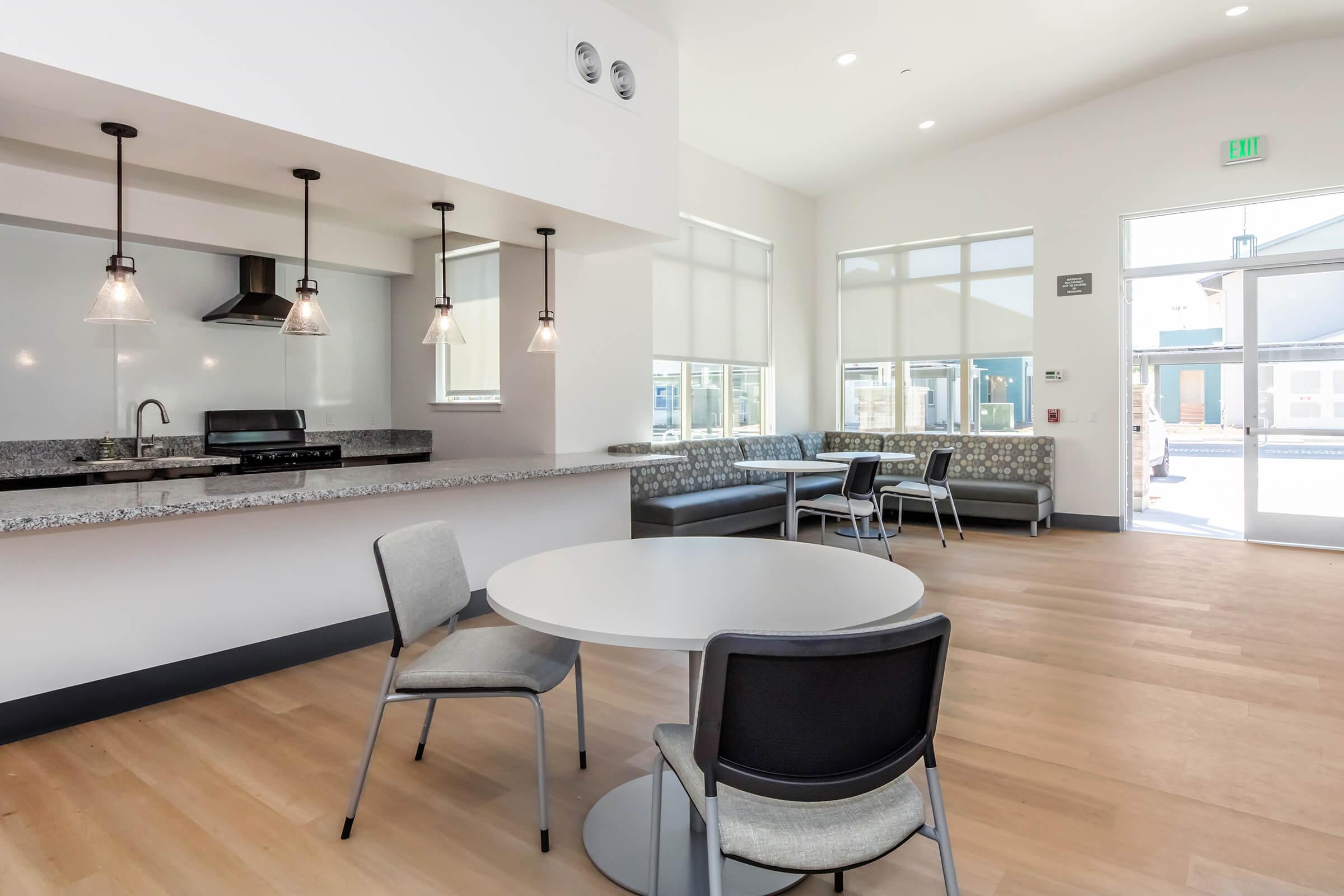 Modern kitchen and dining area featuring a round table with chairs, a gray bench along one wall, and pendant lights hanging above a countertop. Large windows provide natural light, and there is a doorway leading outside. The design is clean and contemporary, with a focus on a spacious layout.