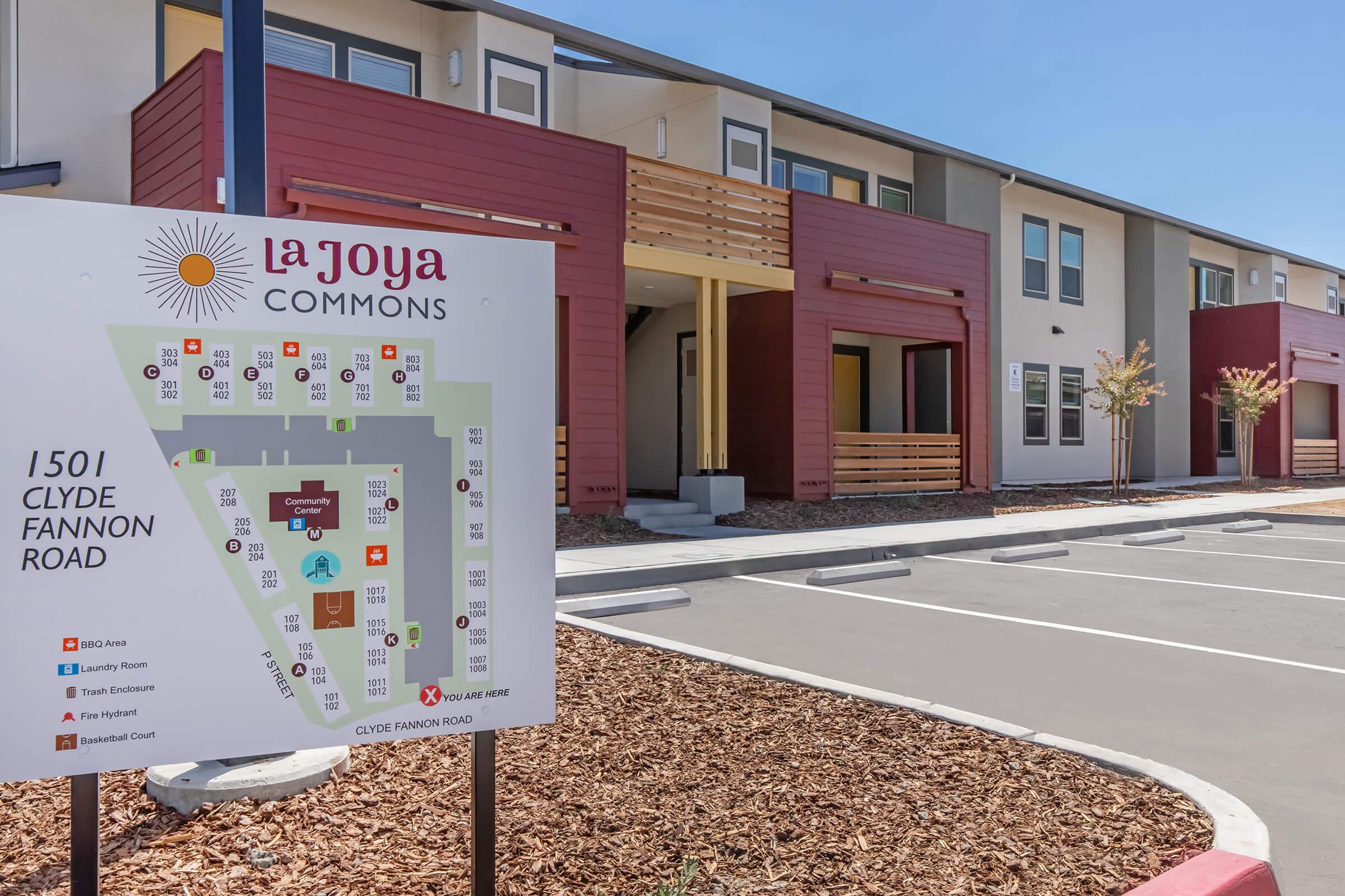 Sign at La Joya Commons displaying a layout of the property and nearby amenities. The building features a modern design with red and beige exteriors and a landscaped area with mulch. Parking spaces are visible in front of the building, and trees provide greenery around the entrance.