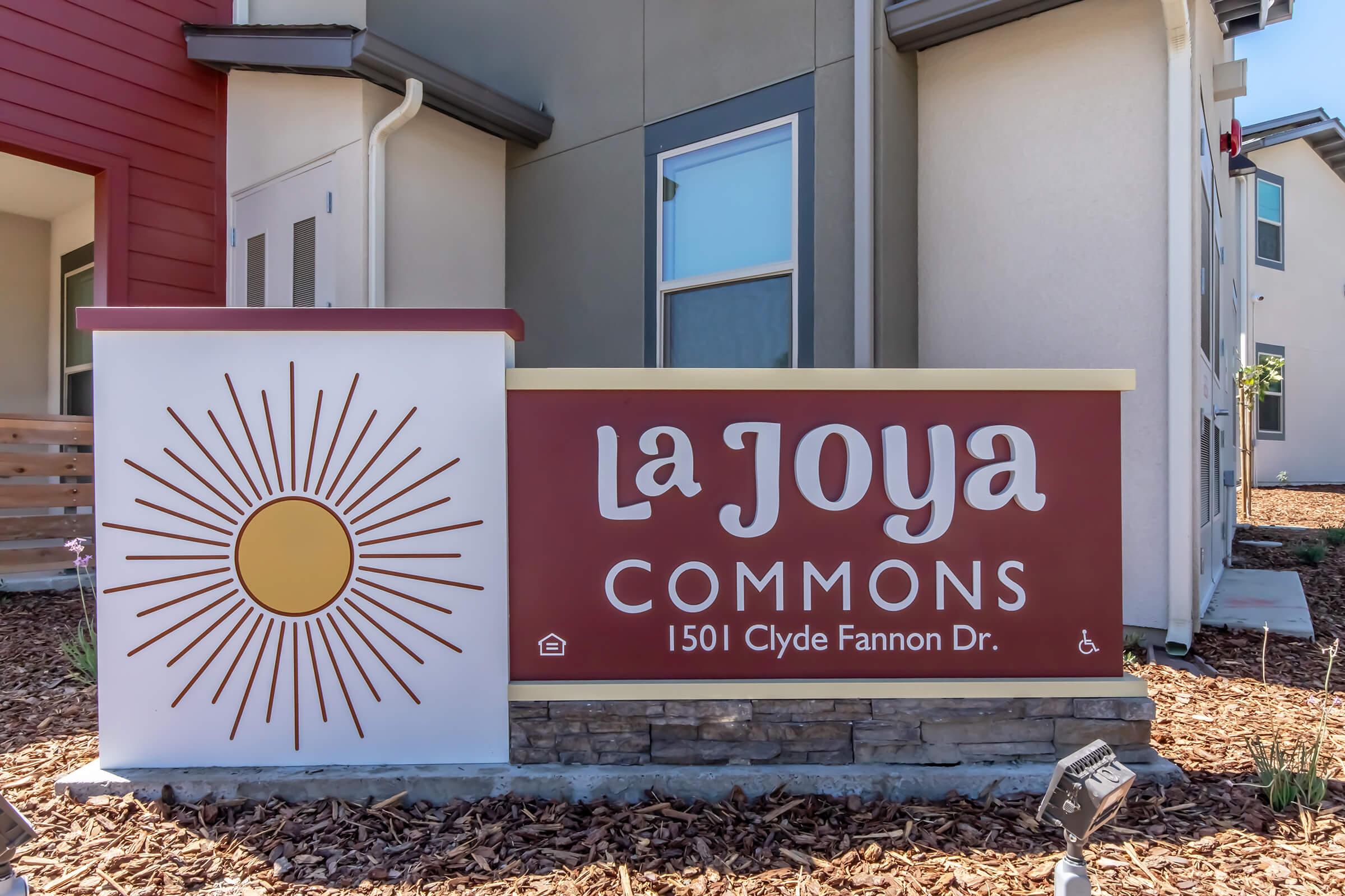 Sign for La Joya Commons featuring a sun graphic, with the address "1501 Clyde Fannon Dr." The sign uses a bold font in warm colors, set against a modern building backdrop. Decorative landscaping surrounds the sign, enhancing the welcoming appearance of the property.