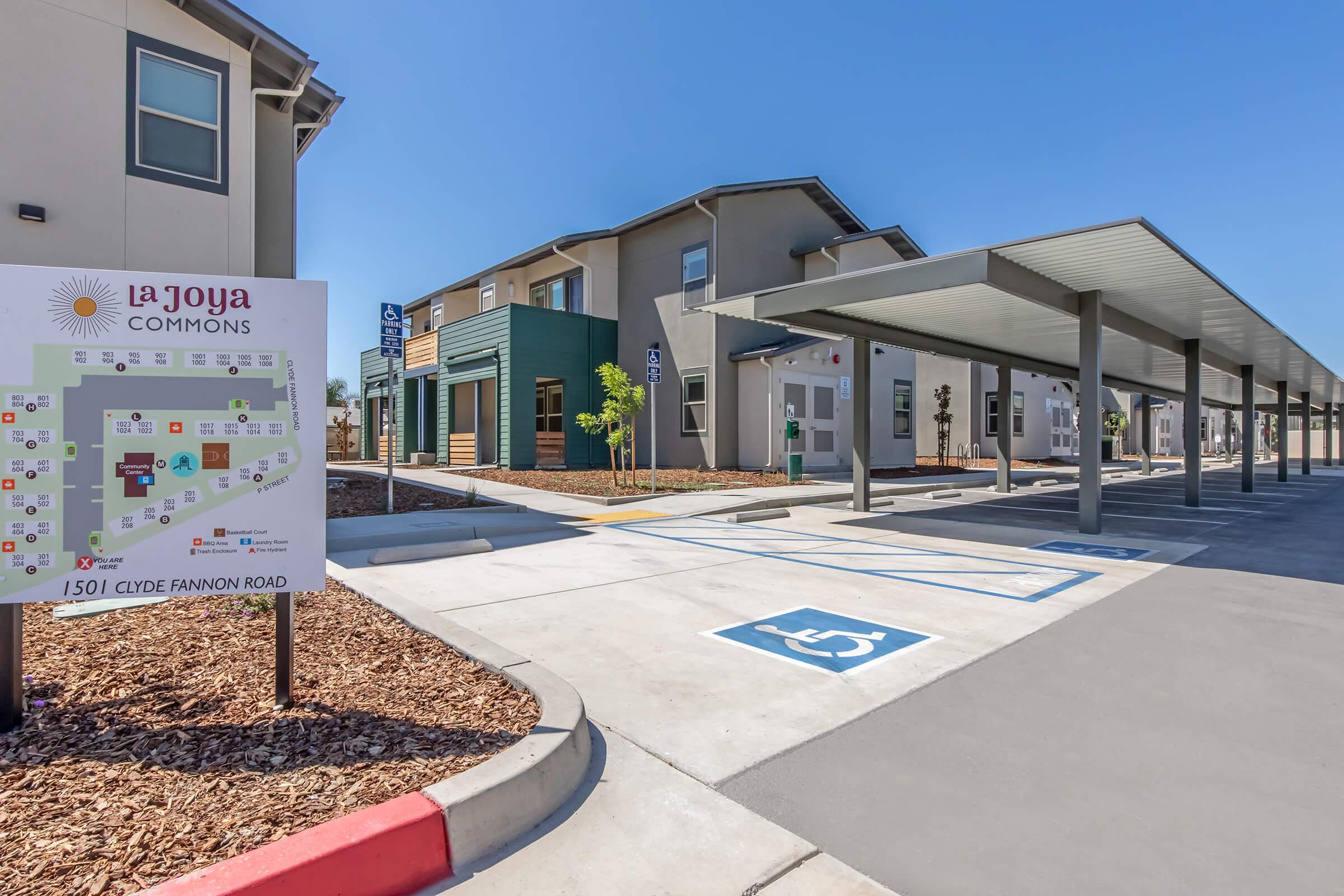 Sign for La Joya Commons with a map indicating various locations within the community. In the background, a modern building with carports and accessible parking spaces is visible. The sky is clear and blue, and the area is landscaped with mulch and decorative plants.