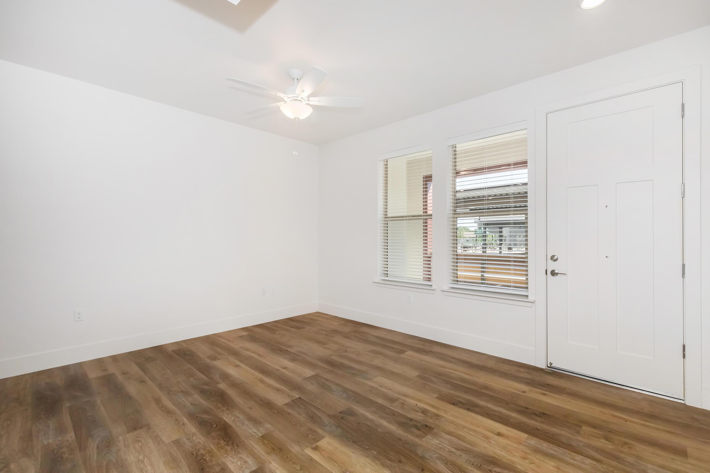 A spacious, empty room with light-colored walls and wood laminate flooring. There is a ceiling fan, two windows with blinds allowing natural light, and a white door. The room appears bright and inviting, perfect for customization or furnishing.