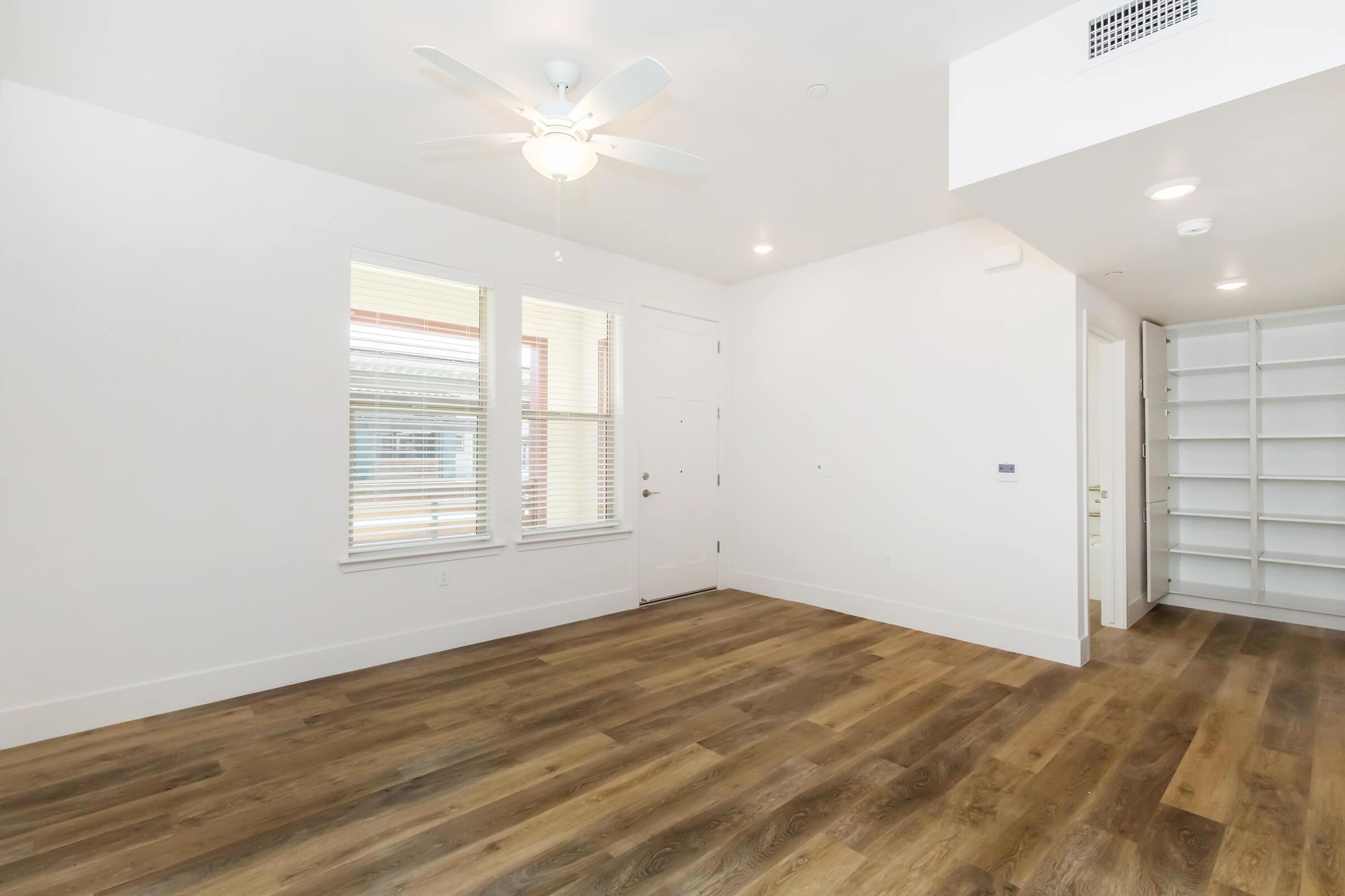A bright, spacious room with light-colored walls, a ceiling fan, and wooden flooring. There are large windows allowing natural light, a front door, and a closet with white shelving. The room appears empty and ready for furnishing, creating an open and inviting atmosphere.
