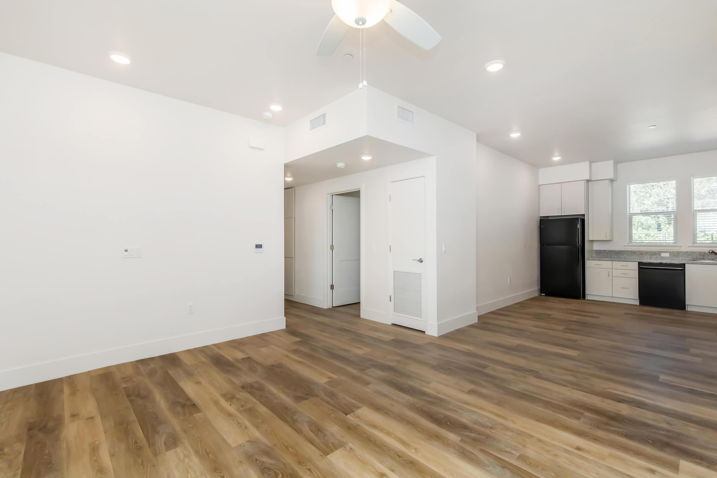 Open living space with light-colored walls and hardwood flooring, featuring a ceiling fan. An entrance leads to a kitchen with modern appliances, including a refrigerator and dishwasher. Natural light streams through windows, enhancing the airy atmosphere.