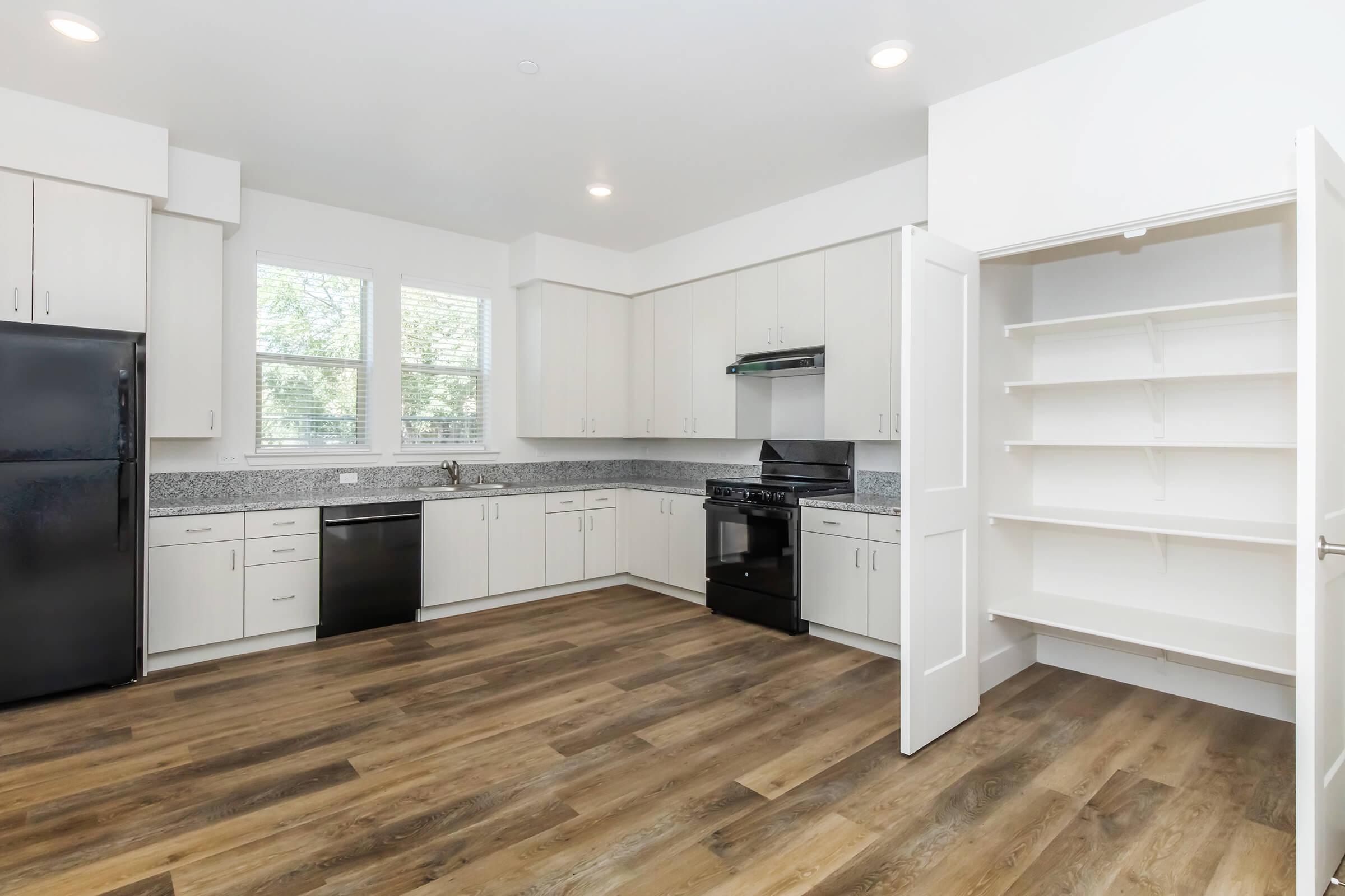 Modern kitchen featuring white cabinets, black appliances including a refrigerator and stove, with a granite countertop. The space has large windows allowing natural light, and a pantry with shelves is visible on the right. The flooring is hardwood, giving the kitchen a warm and inviting appearance.