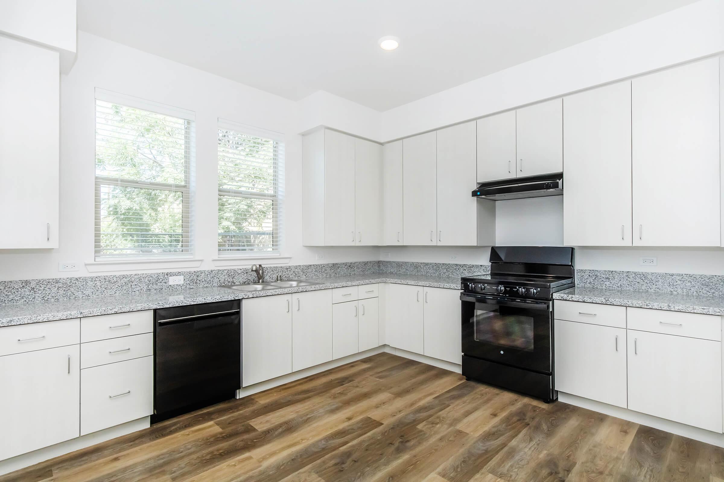 A modern kitchen featuring white cabinets, a black stove and dishwasher, and a granite countertop. Natural light enters through two large windows, illuminating the space with a light wood floor. The design is clean and contemporary, ideal for cooking and entertaining.