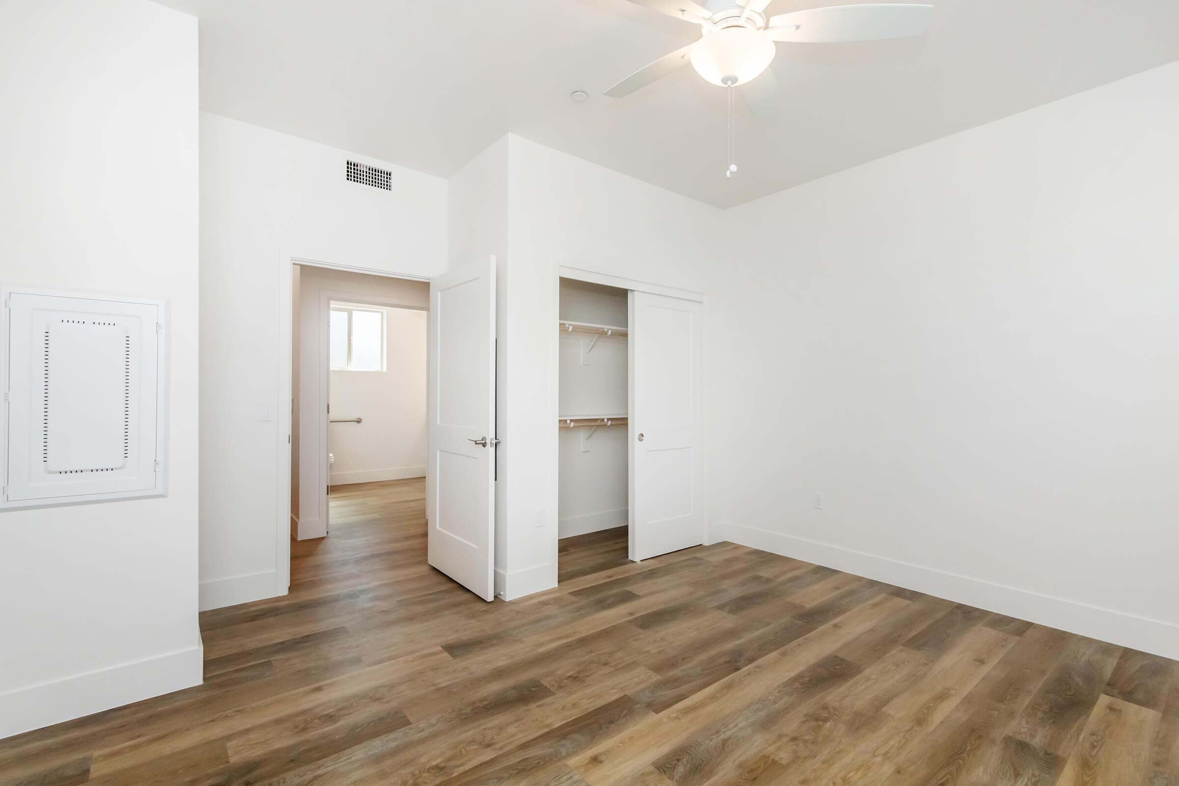 A bright, empty bedroom with light wood flooring and white walls. It features a ceiling fan, a closet with sliding doors, and an open doorway leading to another room. The space is uncluttered, creating a clean and modern aesthetic. Natural light enters through a window in the adjacent room.