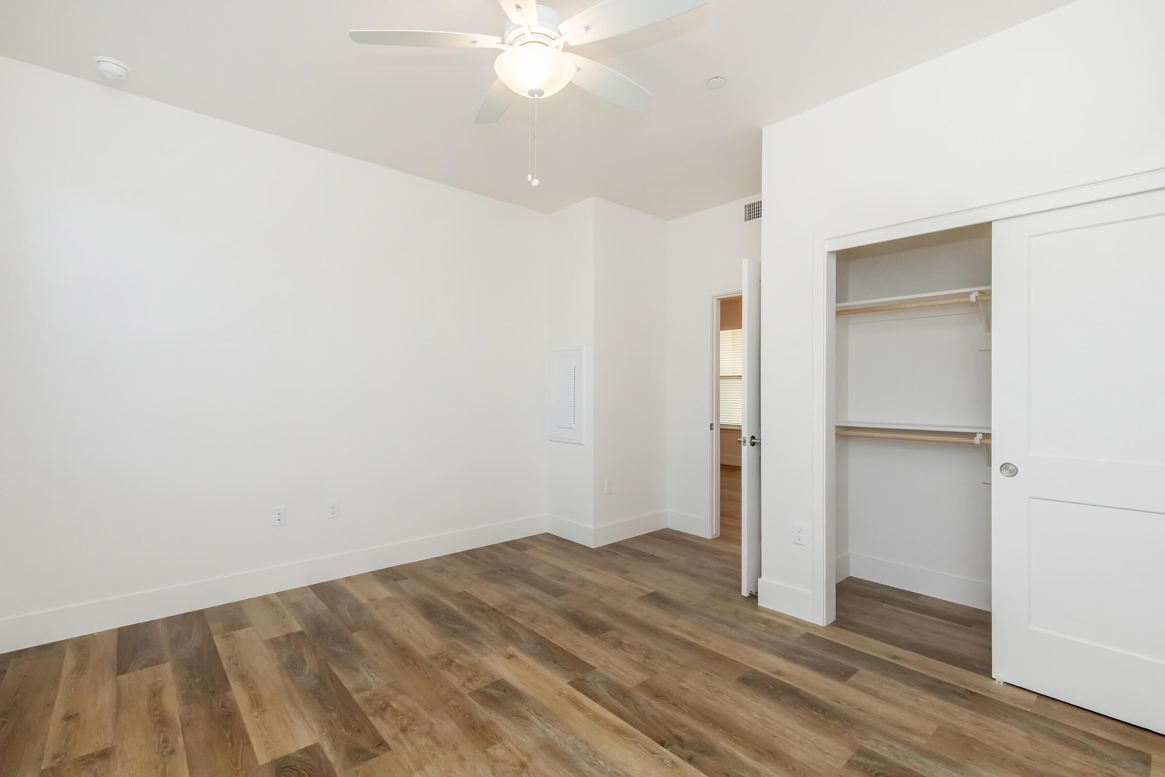 A bright, minimalistic bedroom featuring light-colored walls, a ceiling fan, and hardwood flooring. The room includes a closet with open shelving and a door leading to a bathroom area in the background. Natural light illuminates the space, creating a welcoming atmosphere.