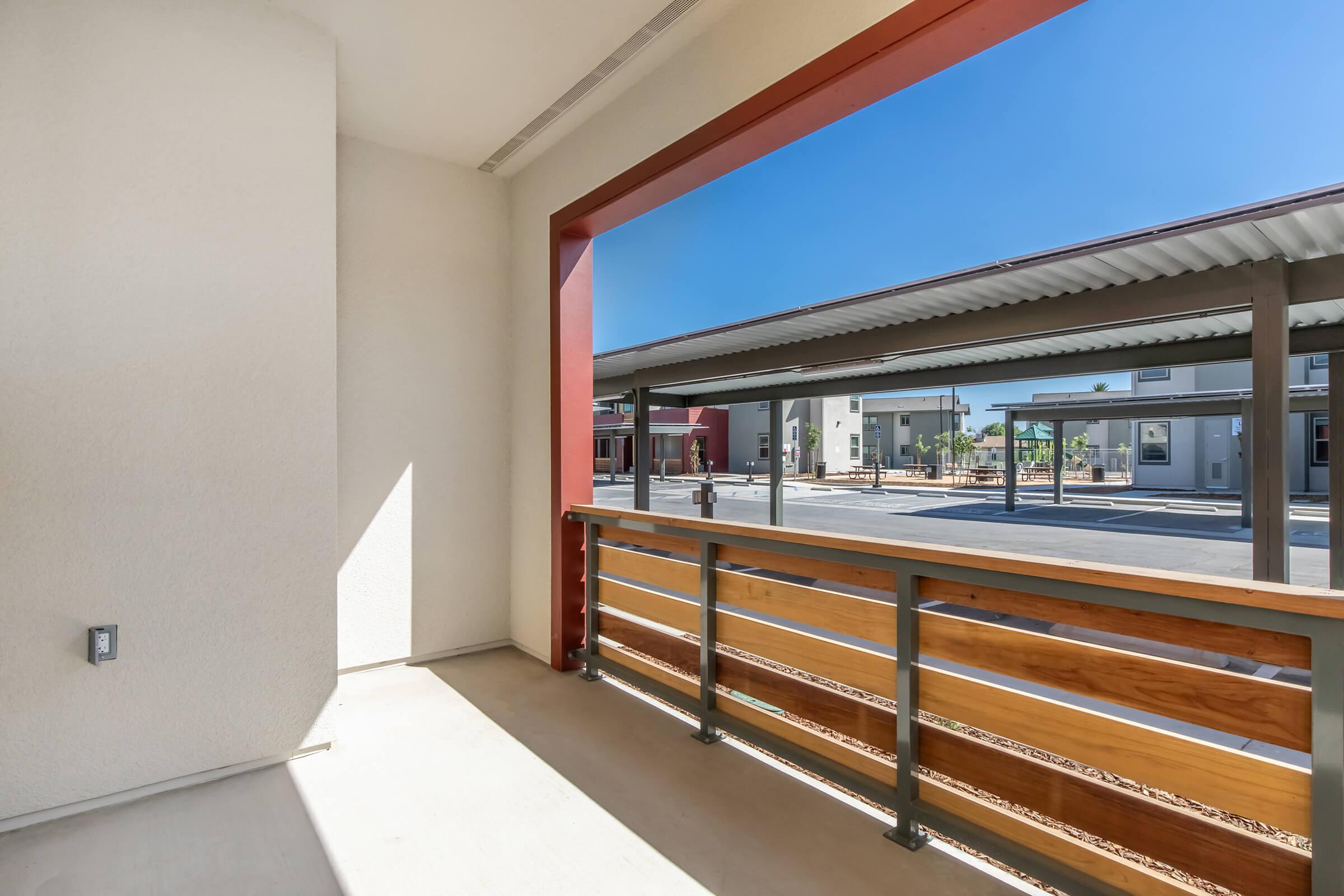 View from a balcony showing a spacious outdoor area with red and wooden railings. In the background, there are several residential buildings under clear blue skies. The setting appears modern and well-lit, highlighting a vibrant community atmosphere.