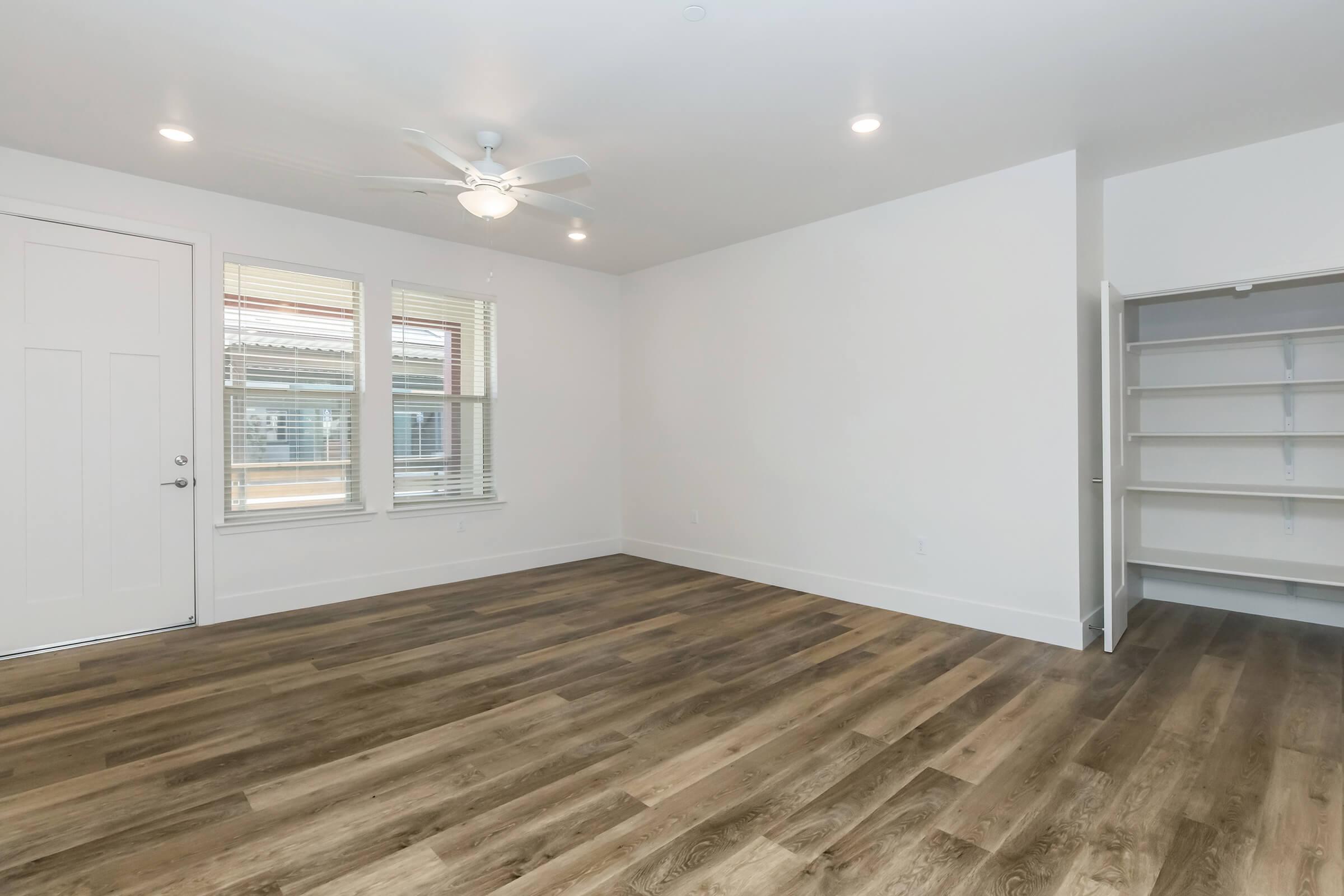 A spacious, empty room with light-colored walls and wooden flooring. It features two windows with blinds, a ceiling fan, and a door leading outside. There is also a built-in shelf unit against one wall, providing ample storage space. Natural light enters the room, creating a bright and inviting atmosphere.