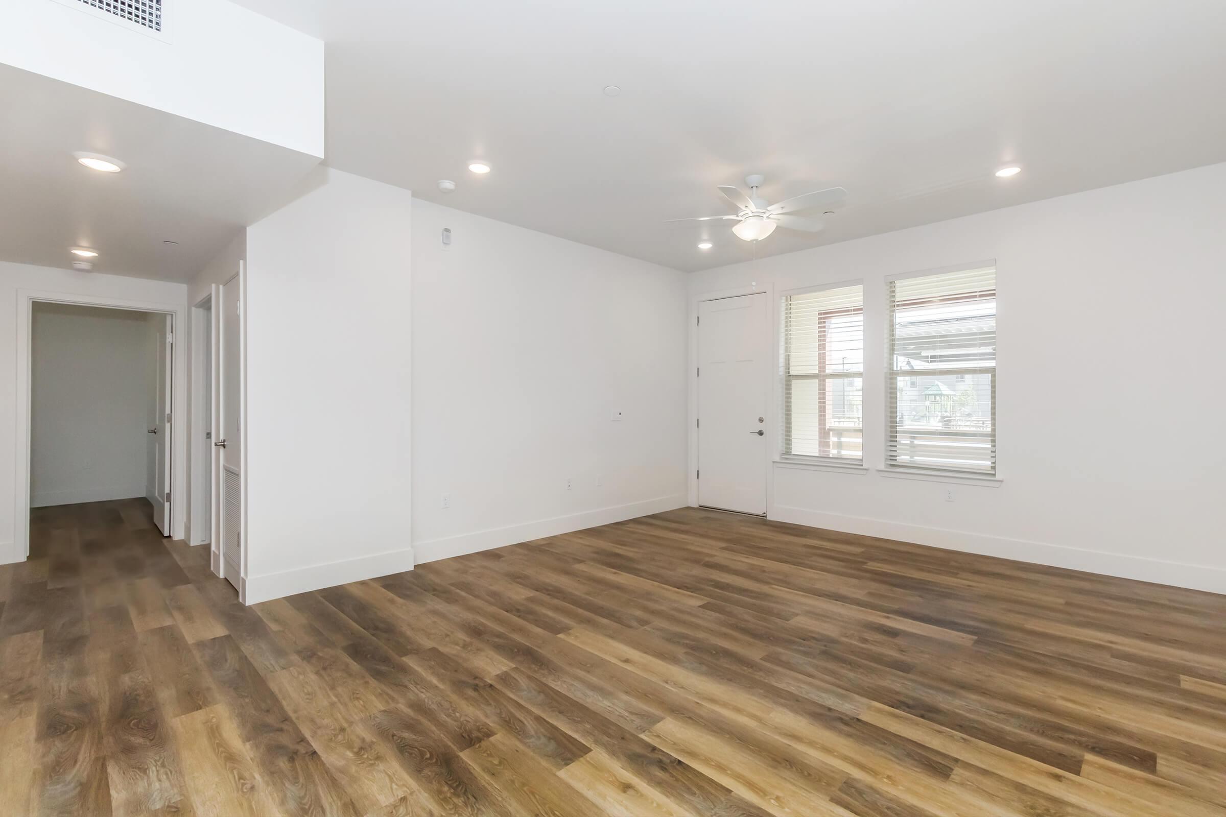 Spacious interior of a modern living room featuring light-colored walls, large windows with blinds, and a ceiling fan. The floor is made of warm-toned wooden planks, and there are doors leading to other rooms visible in the background. The room is well-lit and inviting.