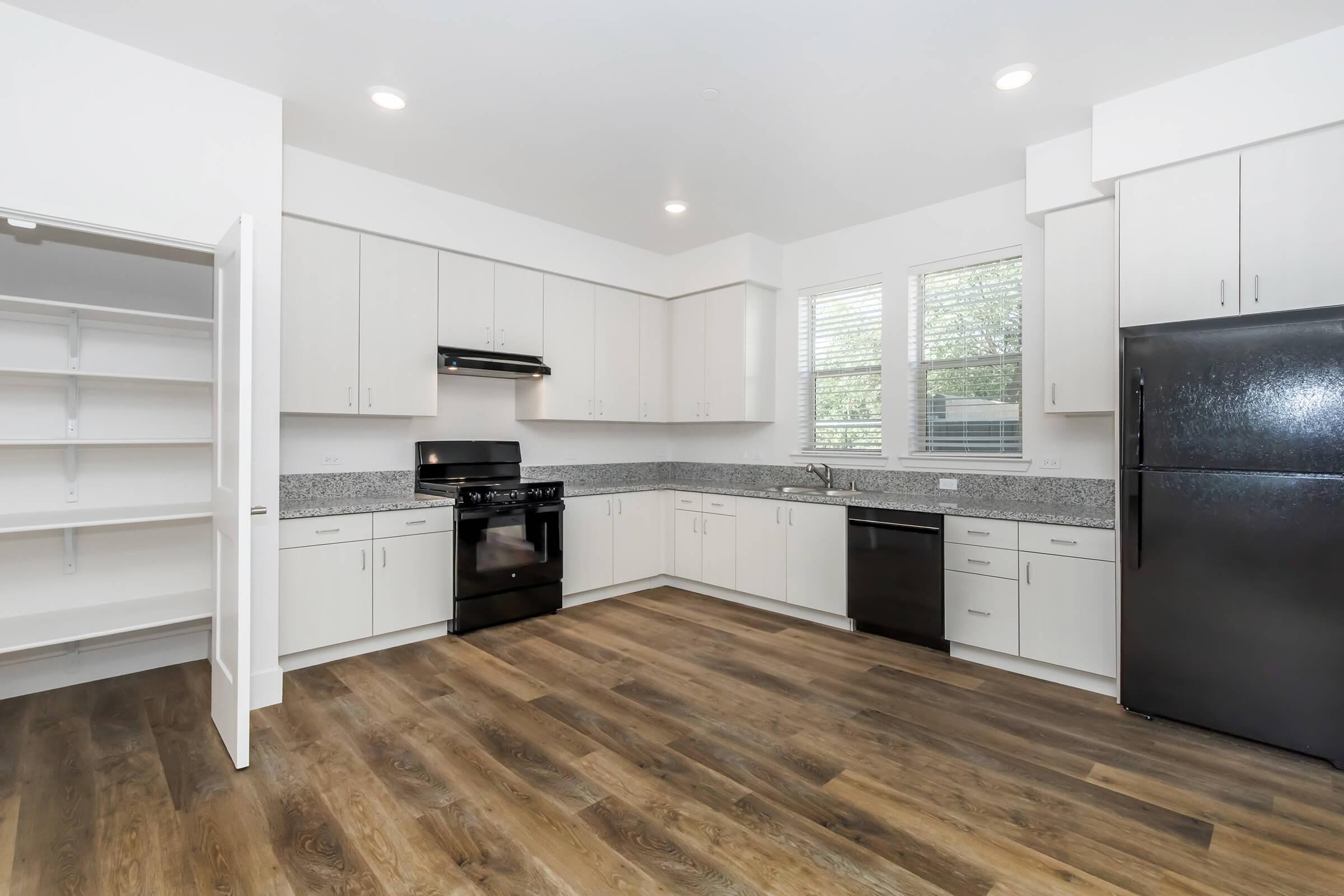 Modern kitchen featuring white cabinetry, black appliances, and a spacious layout. It includes a black refrigerator, stove, and dishwasher, complemented by a countertop with a patterned backsplash. The room has large windows for natural light and a pantry with open shelving. The floor is made of wood-like material.