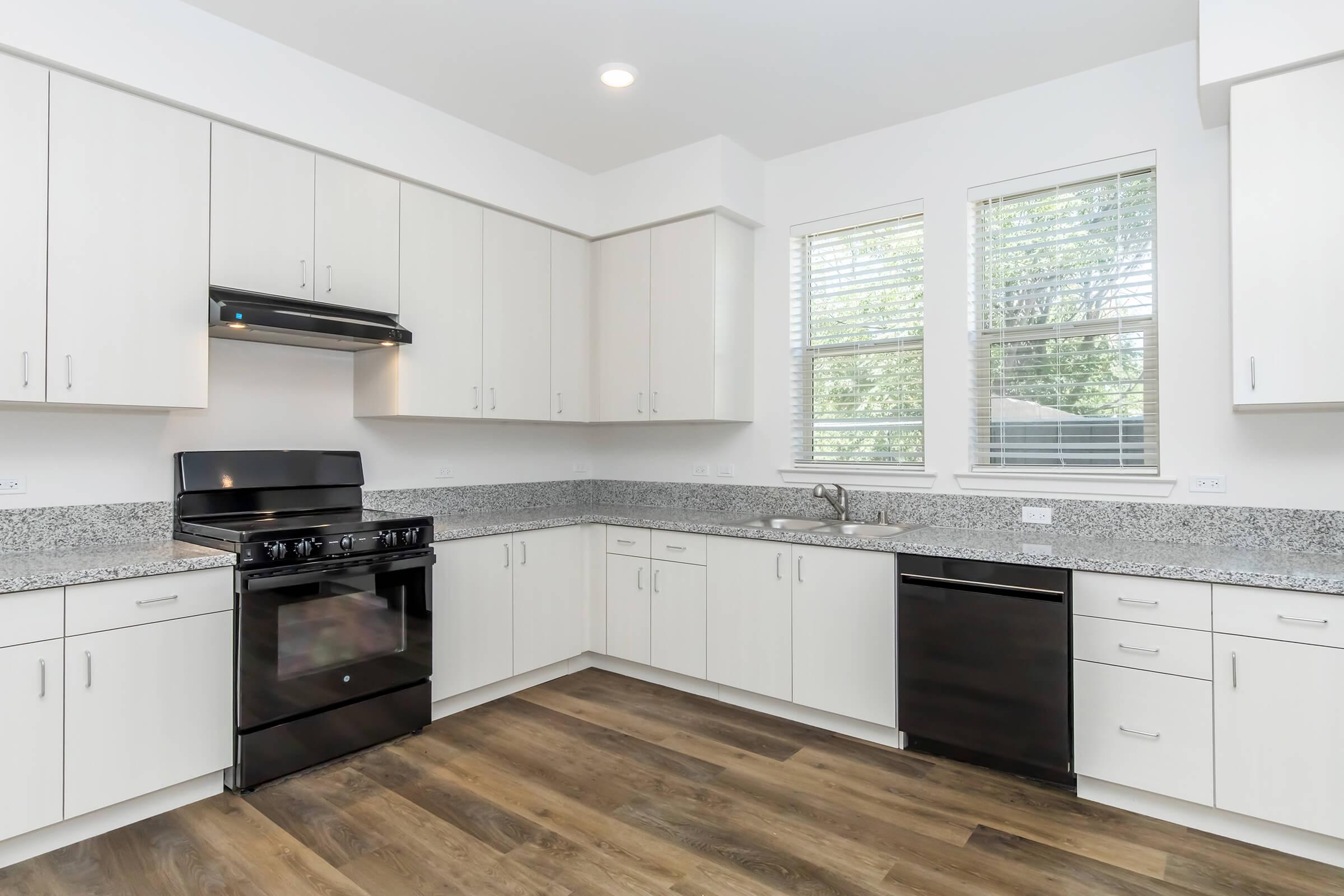 Modern kitchen with a spacious layout featuring white cabinetry, dark countertops, and stainless steel appliances. The kitchen includes a black gas stove, a dishwasher, and a sink under a window, allowing natural light to brighten the space. The floor is made of wooden planks, enhancing the overall aesthetic.