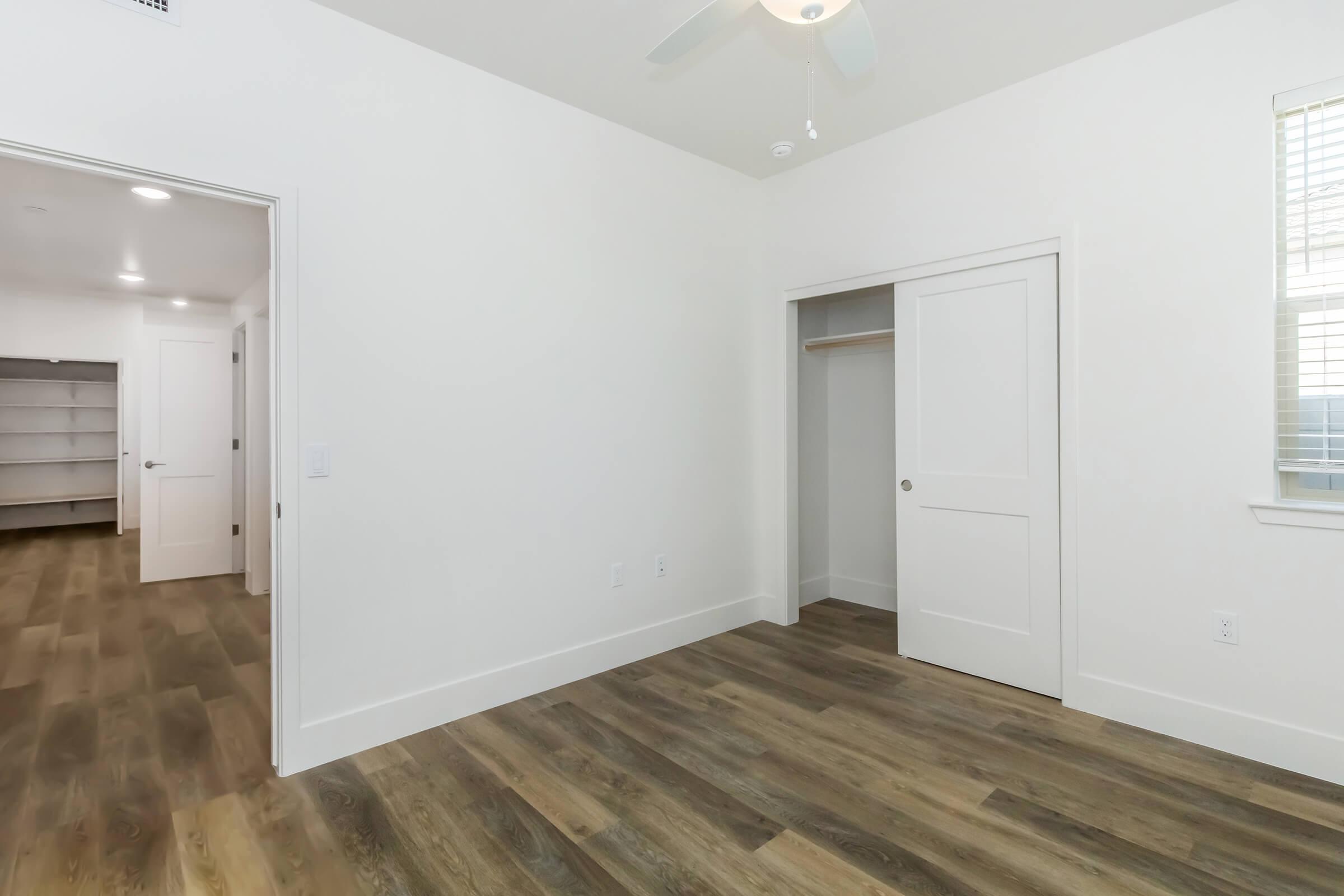 A clean, minimalistic room with light-colored walls and wood-style flooring. A ceiling fan is visible, and there is an open door leading to a closet. Natural light streams in through a window, illuminating the space. The opposite wall features another door leading to a different area, giving a sense of openness.