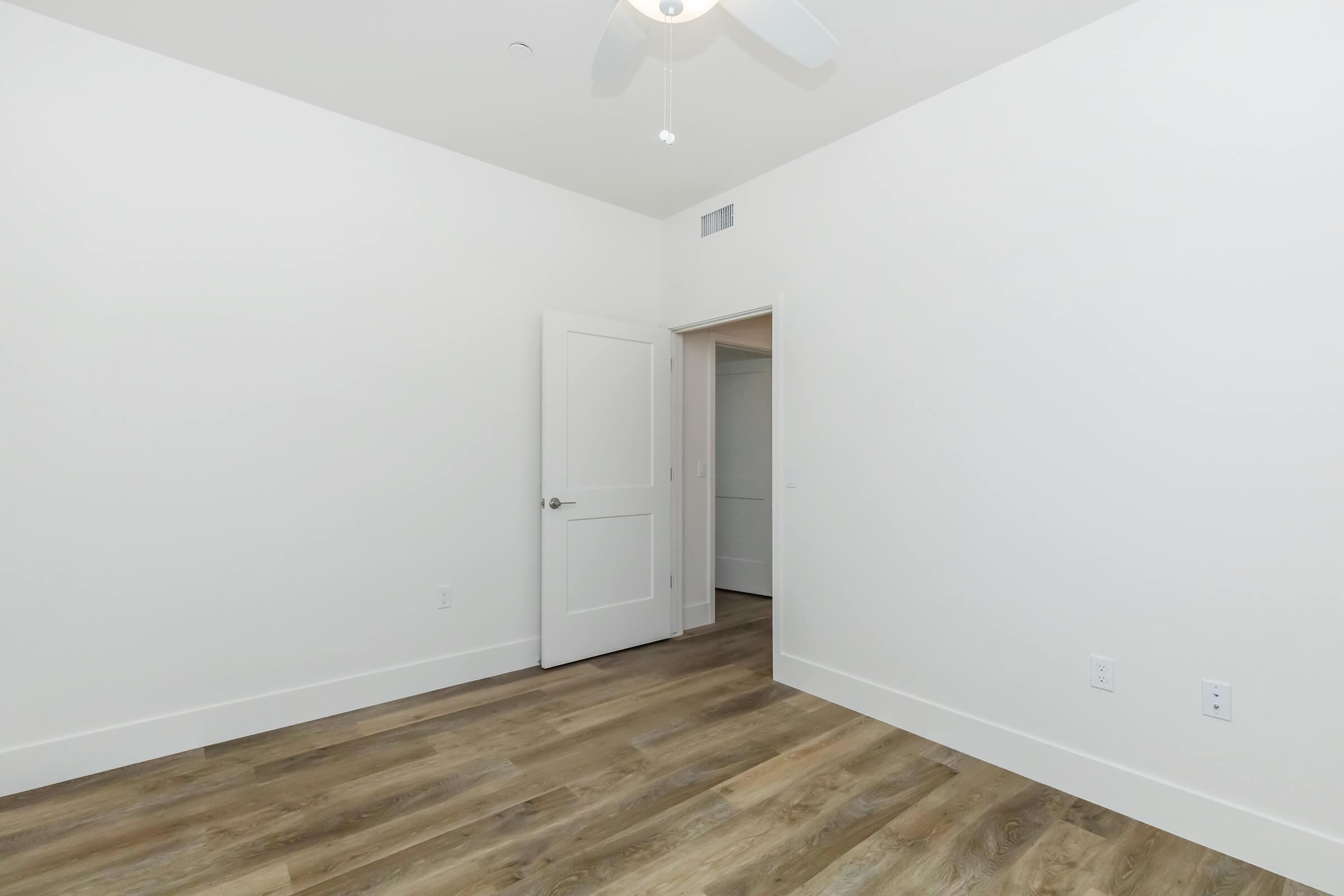Empty room featuring light-colored walls, a wooden floor, and a ceiling fan. A white door is slightly ajar, leading to another room. The overall atmosphere is clean and minimalist, suitable for various uses.