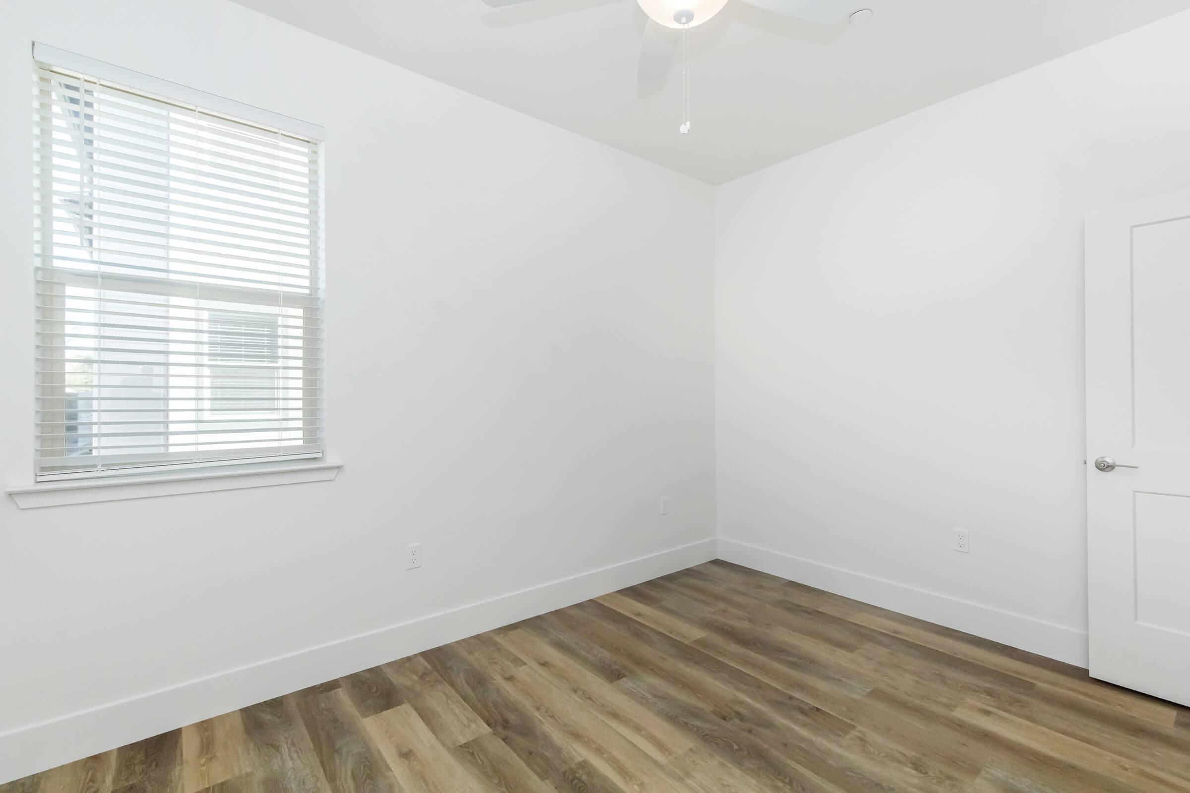 A bright, empty room with white walls and a ceiling fan. There is a window with blinds allowing natural light to enter, and the flooring is made of wooden planks. The door is closed and has a modern handle, contributing to the clean and minimal aesthetic of the space.