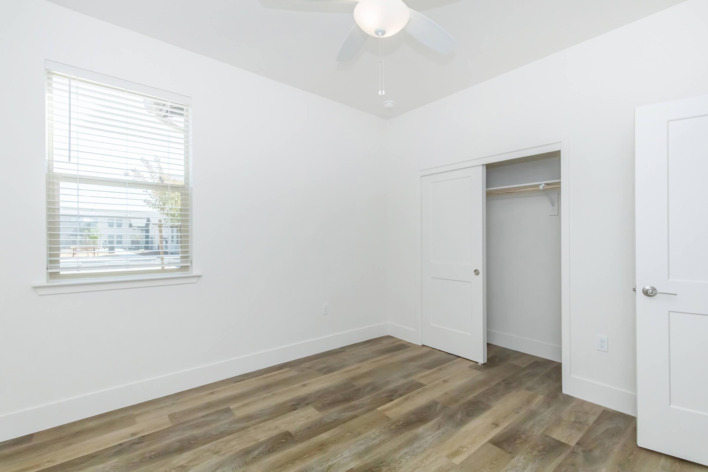 Bright, empty room with light-colored walls and a ceiling fan. A large window with blinds lets in natural light, and there is a closet with sliding doors. The floor features a wood-like design, creating a modern and minimalist atmosphere.