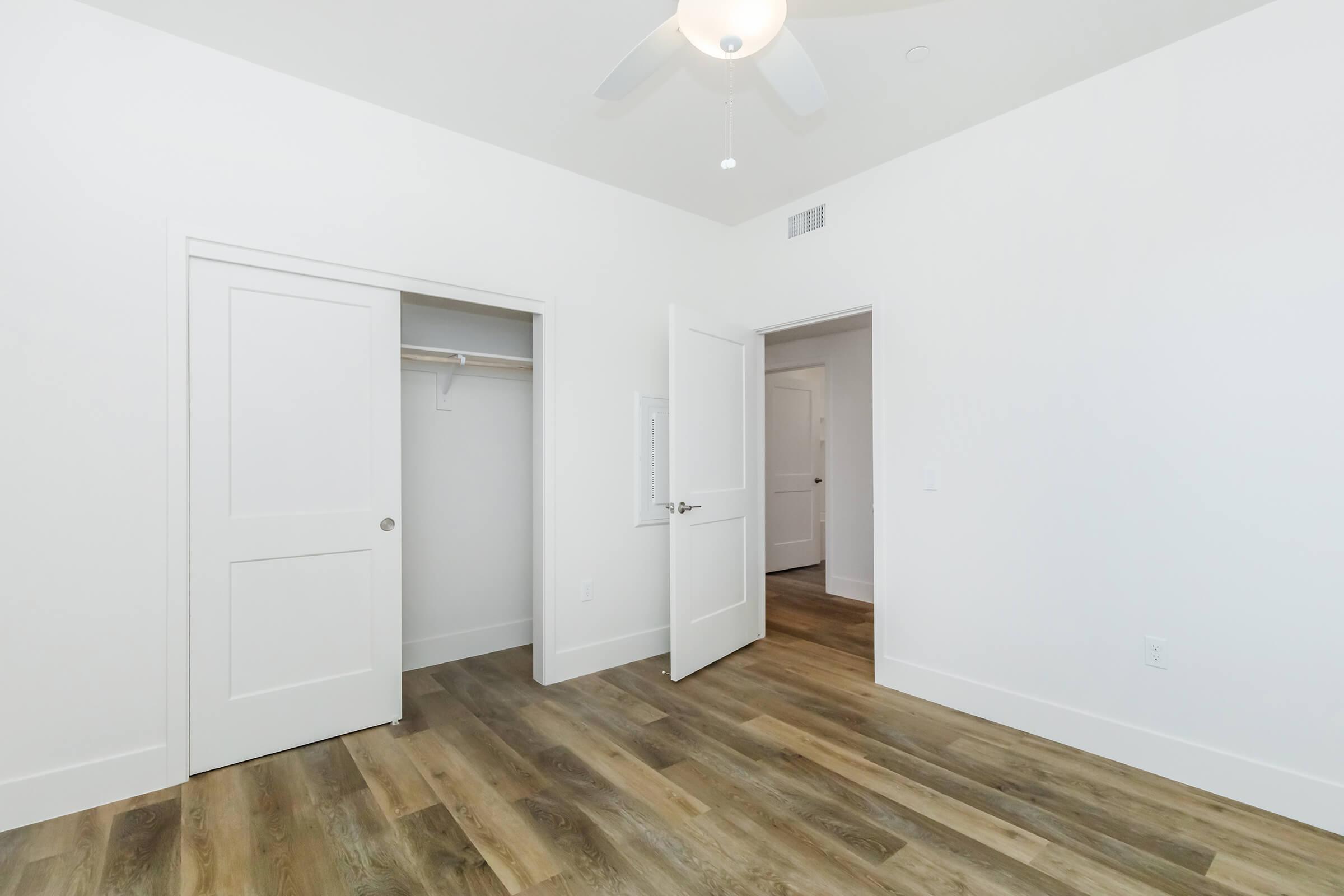 A spacious and bright bedroom featuring light-colored walls, a ceiling fan, and laminate flooring. There are two open doors: one leading to a closet and the other to an adjoining room. Natural light floods the area, creating a welcoming atmosphere.