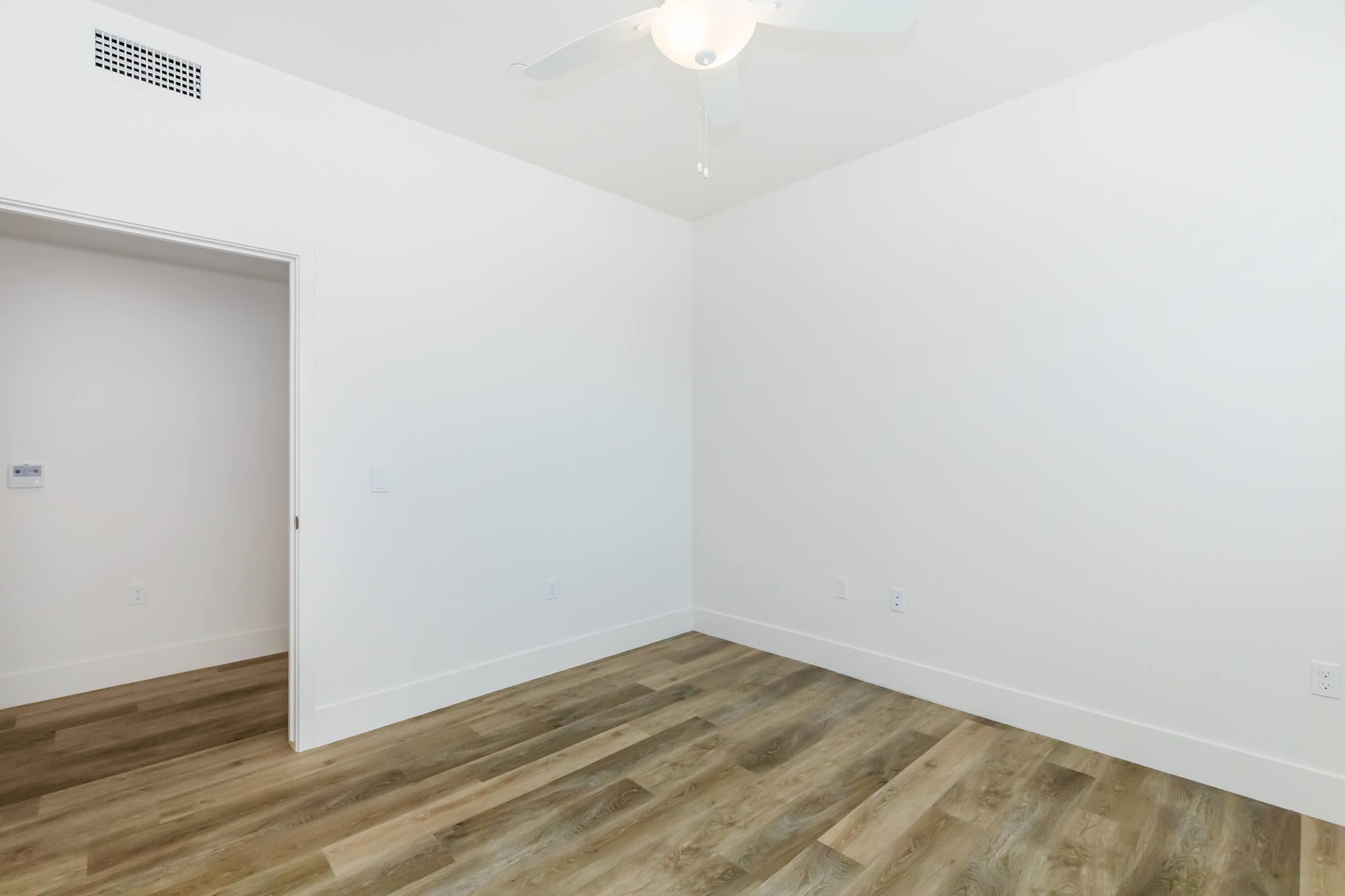 A spacious, empty room with light-colored walls and a ceiling fan. The floor is made of light wood or laminate, and there is an open doorway leading to another room. Natural light illuminates the space, creating a bright and airy atmosphere.