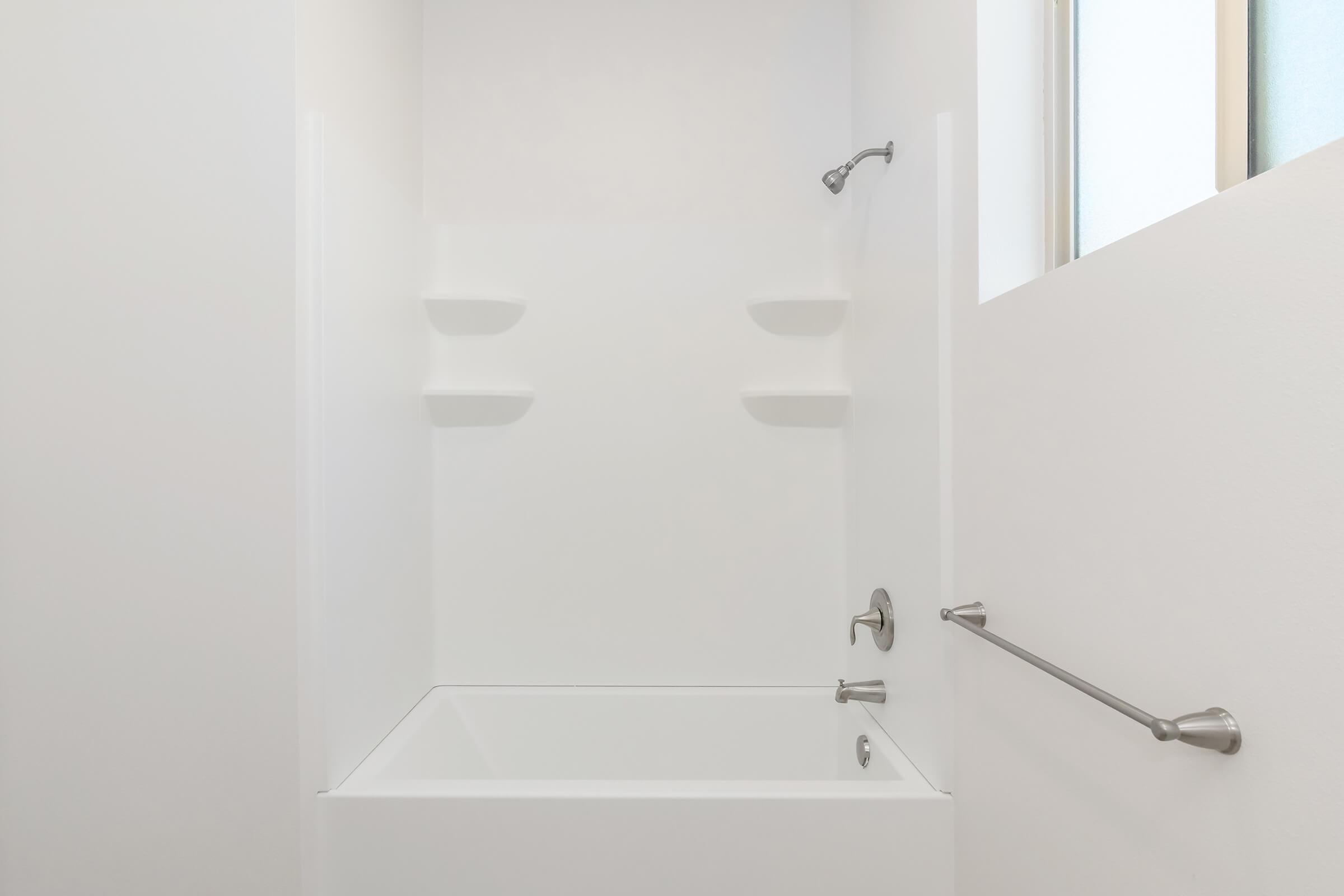 A clean, minimalist bathroom featuring a white bathtub and shower area. The walls are plain and light-colored, with two small shelves incorporated into the design. A grab bar is installed next to the tub for safety, and there is a window allowing natural light into the space.