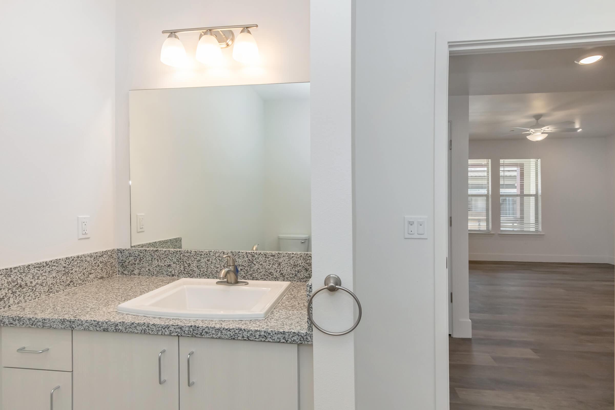 A modern bathroom featuring a granite countertop with a single sink and a large mirror above it. A towel ring is mounted beside the sink. In the background, a room with light-colored walls and hardwood flooring can be seen, showcasing a ceiling fan and window with blinds.