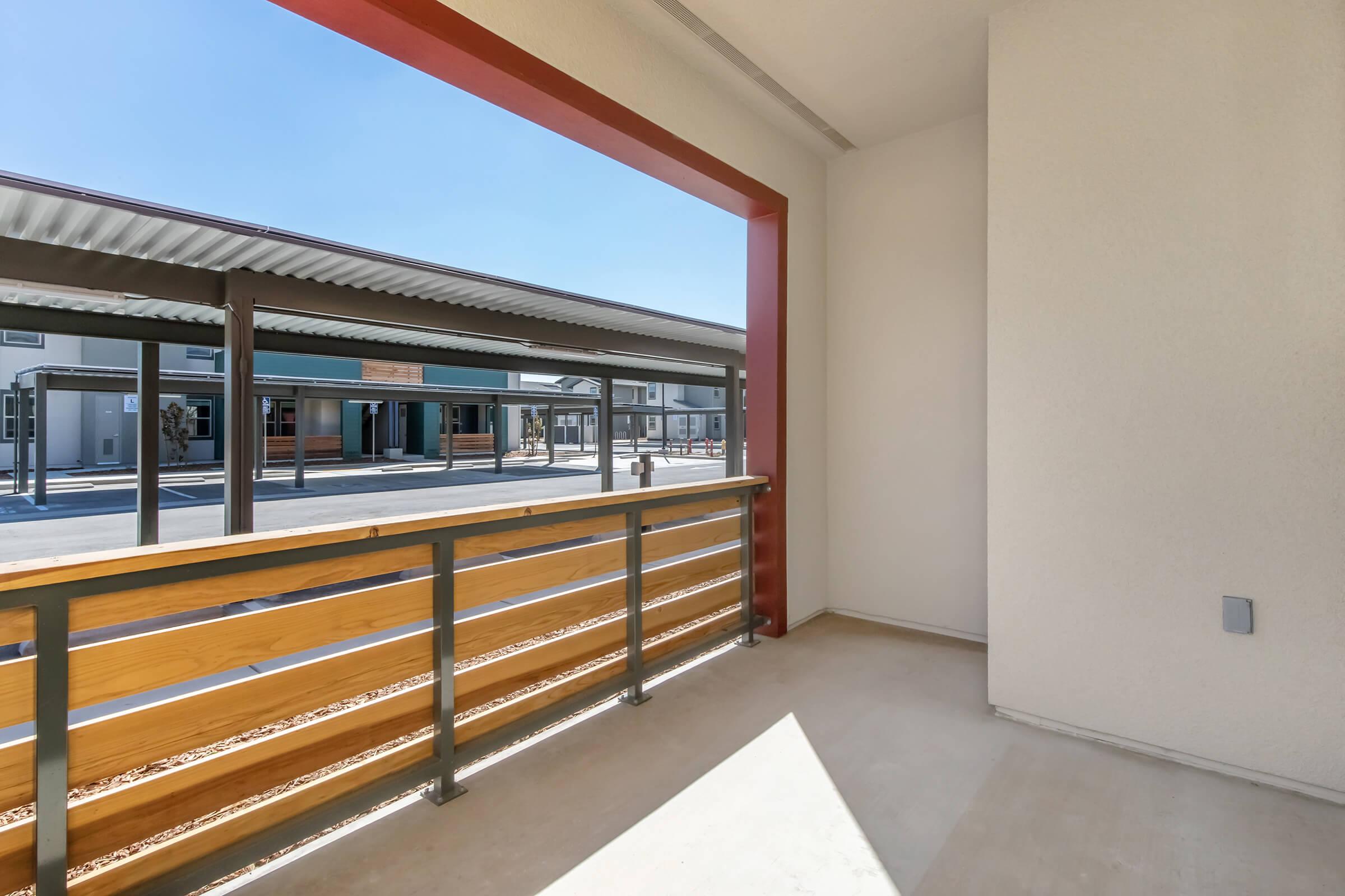 A view of a modern balcony overlooking a courtyard with buildings and walkways. The balcony features a wooden railing and bright sunlight, creating a warm and inviting atmosphere. The background includes structured buildings with awnings and clear blue skies.
