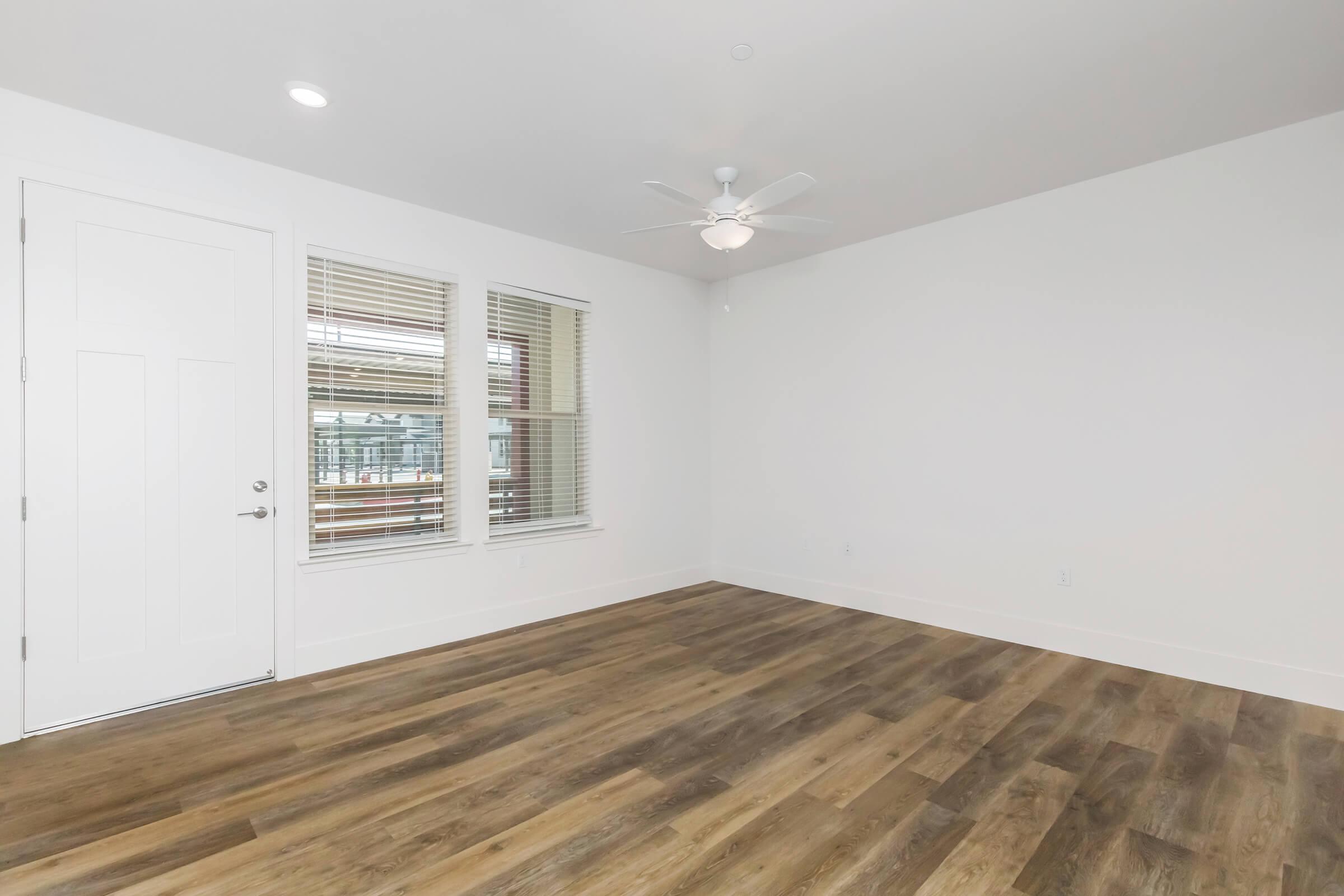 An empty room featuring light-colored walls, hardwood-style flooring, and a ceiling fan. There is a white door on the left and a large window with blinds, allowing natural light to enter the space. The room has a clean and modern aesthetic, with ample space for furniture placement.