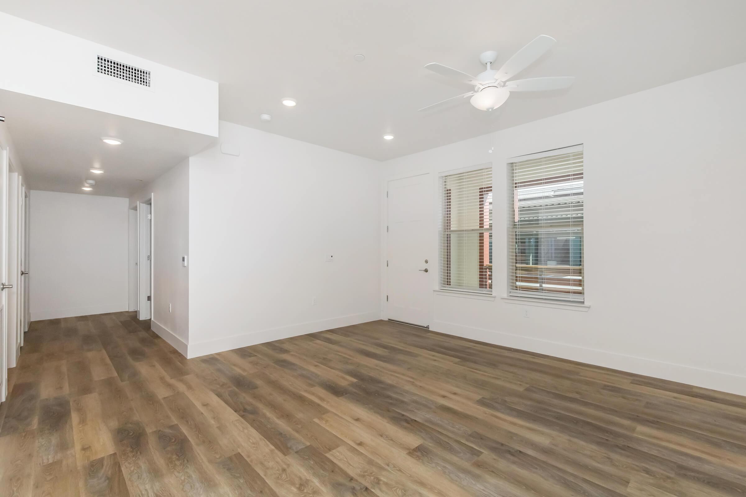 A spacious and well-lit interior of an apartment featuring light-colored walls, large windows, and wooden flooring. The room has a ceiling fan and is free of furniture, creating an open, airy atmosphere. Doorways visible lead to other rooms, suggesting a modern design.