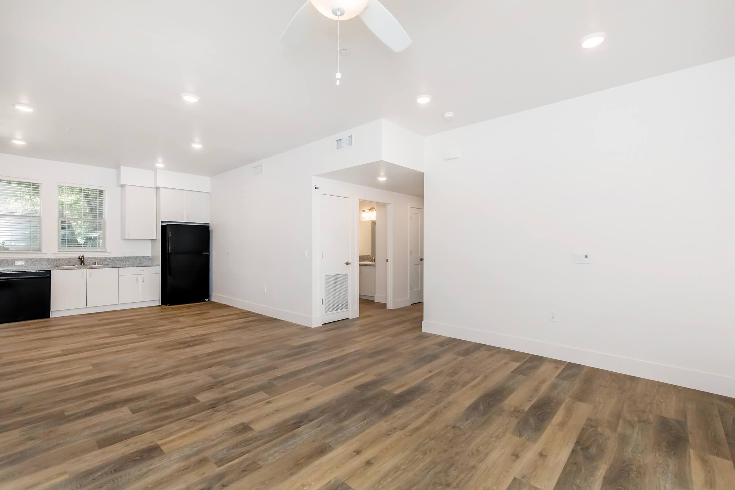 A bright, modern living space featuring polished wooden floors, white walls, and recessed lighting. To the left, a kitchen area with white cabinets and a black refrigerator. A doorway leads to a bathroom, and the space is open and airy, creating a welcoming atmosphere.