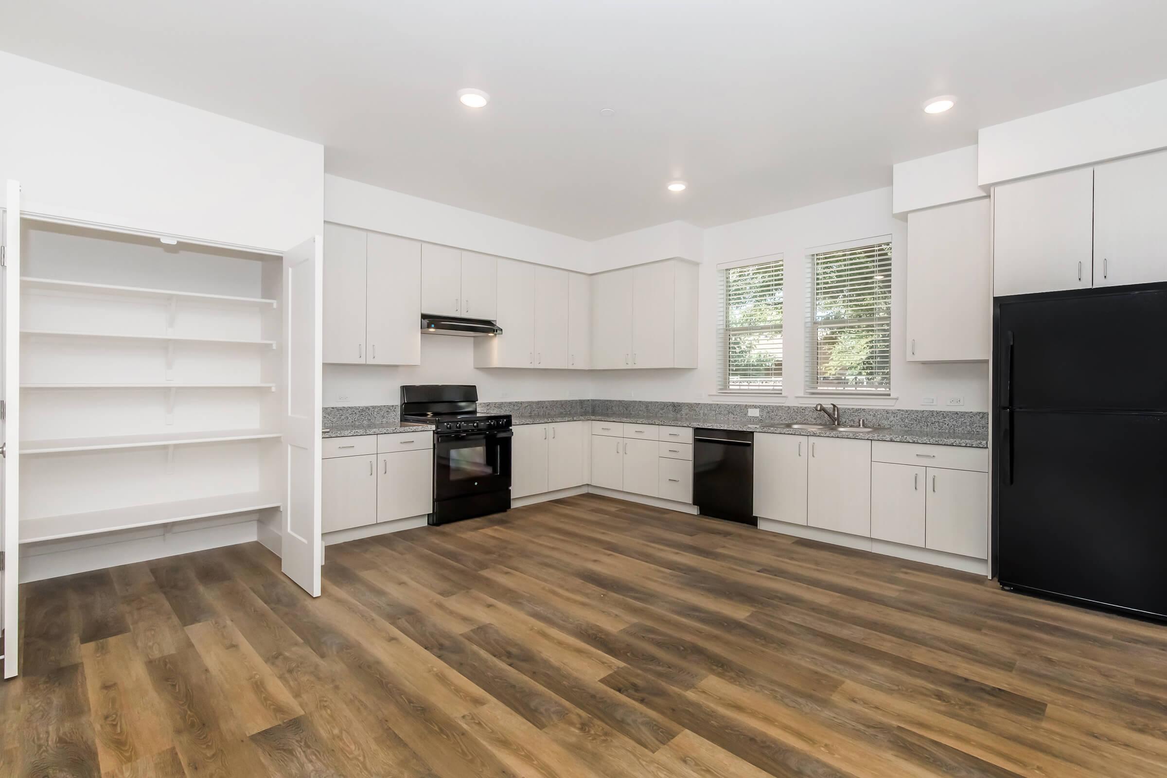 Modern kitchen featuring white cabinetry, granite countertops, black appliances, and a spacious layout. Includes a refrigerator, stove, and ample storage with a pantry. Large windows provide natural light, and the floor is finished with wood-like laminate.