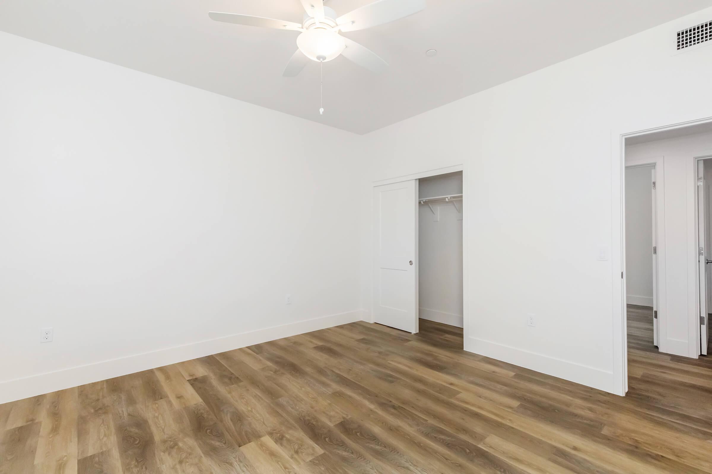 A spacious, empty room featuring light-colored walls, a ceiling fan, and hardwood flooring. There's a closet door on the left and an entrance to another room visible on the right. The overall aesthetic is clean and modern, with ample natural light.