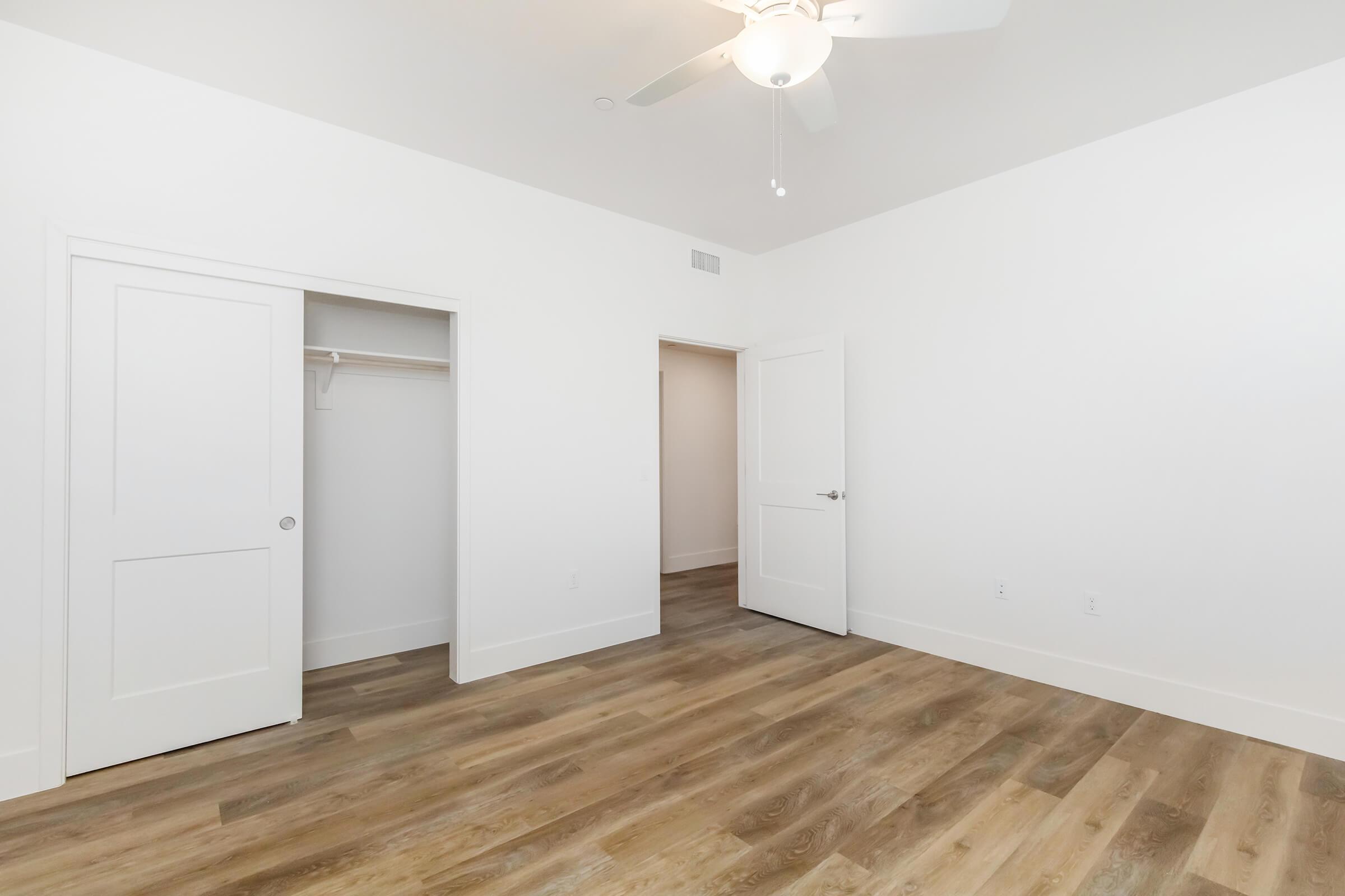 A bright, minimally decorated room featuring light-colored walls and laminate flooring. There are two white doors: one leads to a closet and the other to an adjoining area. A ceiling fan is visible, adding to the room's modern design and comfort.